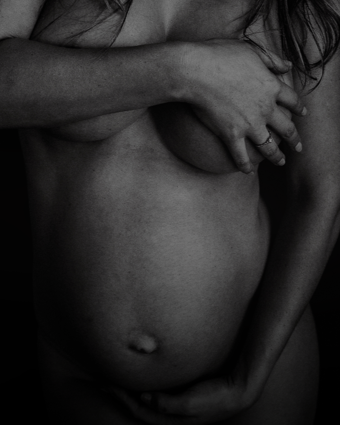 Black-and-white close-up of a pregnant person cradling their bare belly with both hands.