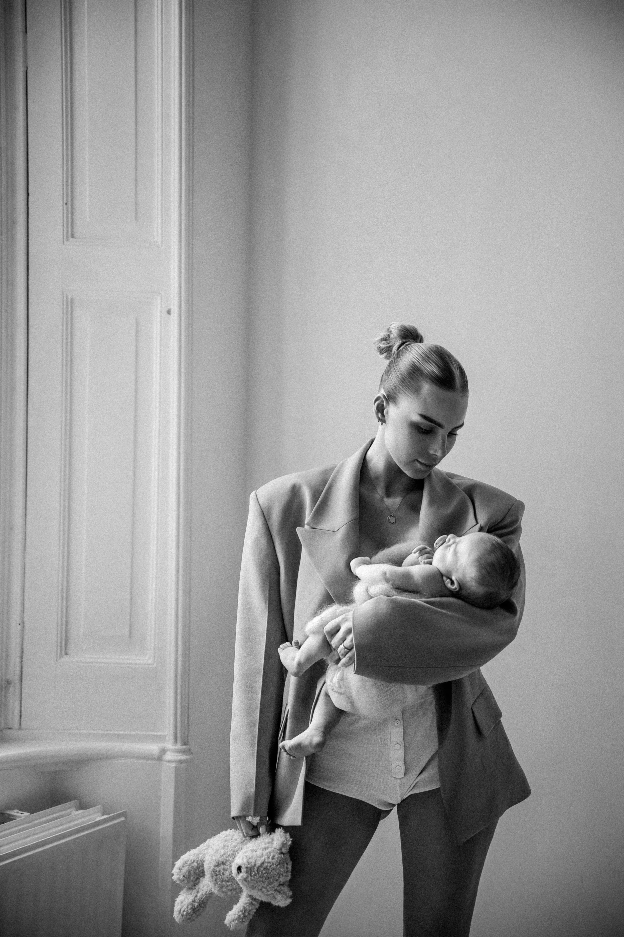 A woman with her hair in a bun holding a baby in her arms. The woman is looking down at the baby, and the baby is looking up at her. The woman is dressed in a blazer, and the baby is dressed in light-colored clothing. The woman is also holding a plush toy dog in her left hand.