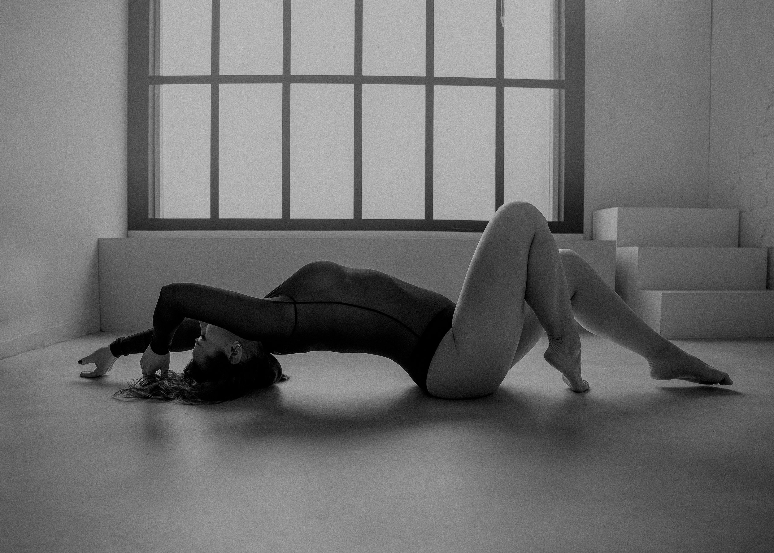 Een vrouw in sportkledij strekt zich uit op de vloer, met haar rug in een boog, haar hoofd en armen achter haar, voor een groot raam in een minimalistische, industriële ruimte.