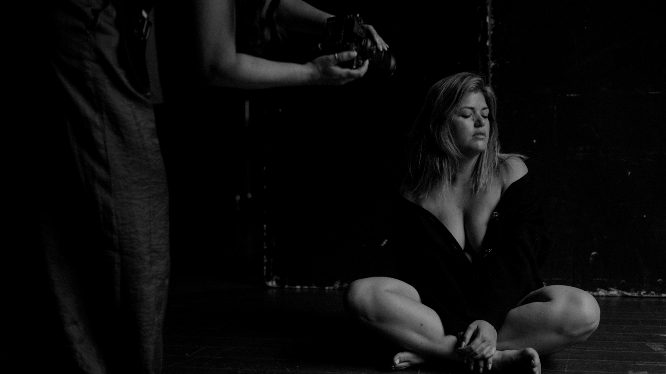 Vrouw met blond haar, zittend op de vloer met gekruiste benen en gesloten ogen, in een zwarte open top — intiem portret in zacht schemerlicht.