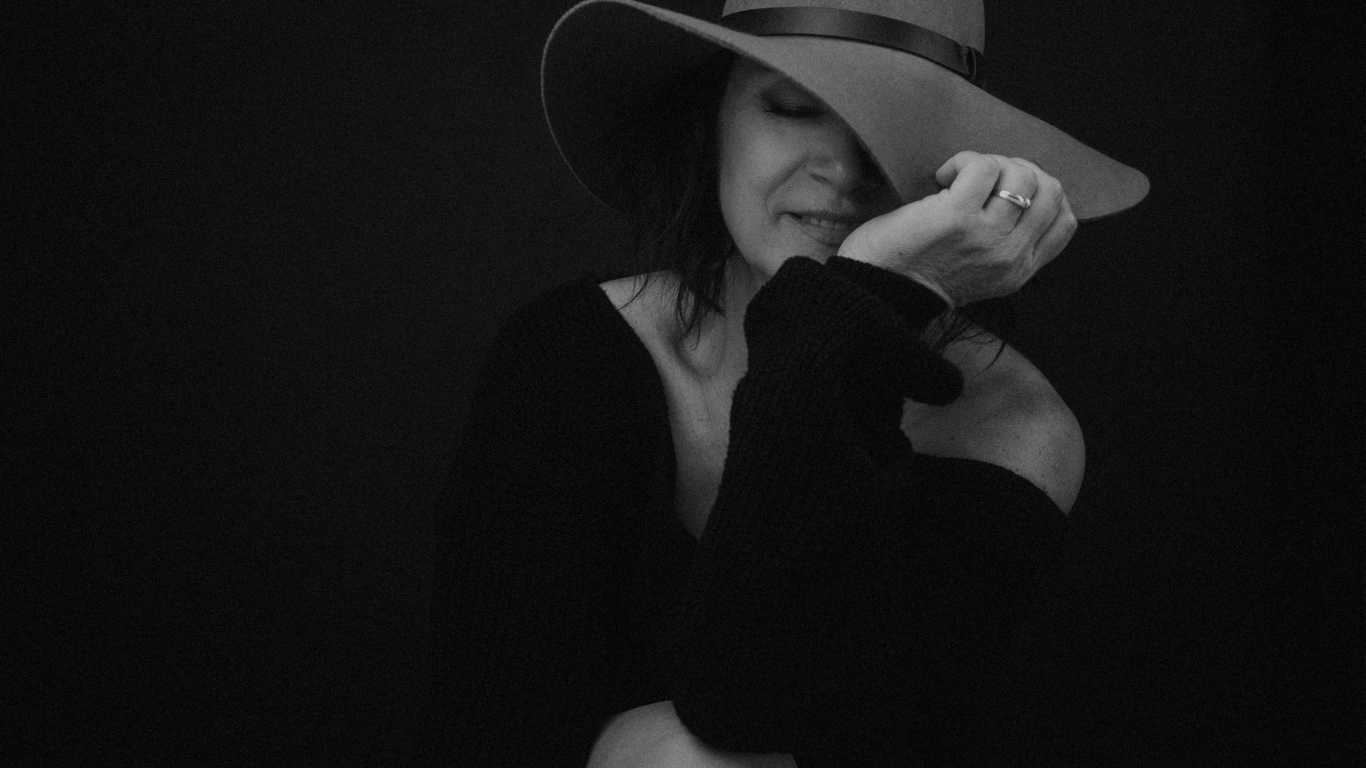 Vrouw met grote hoed die haar gezicht deels bedekt, in een donkere outfit tegen een donkere achtergrond — een ingetogen, mysterieus boudoirportret.