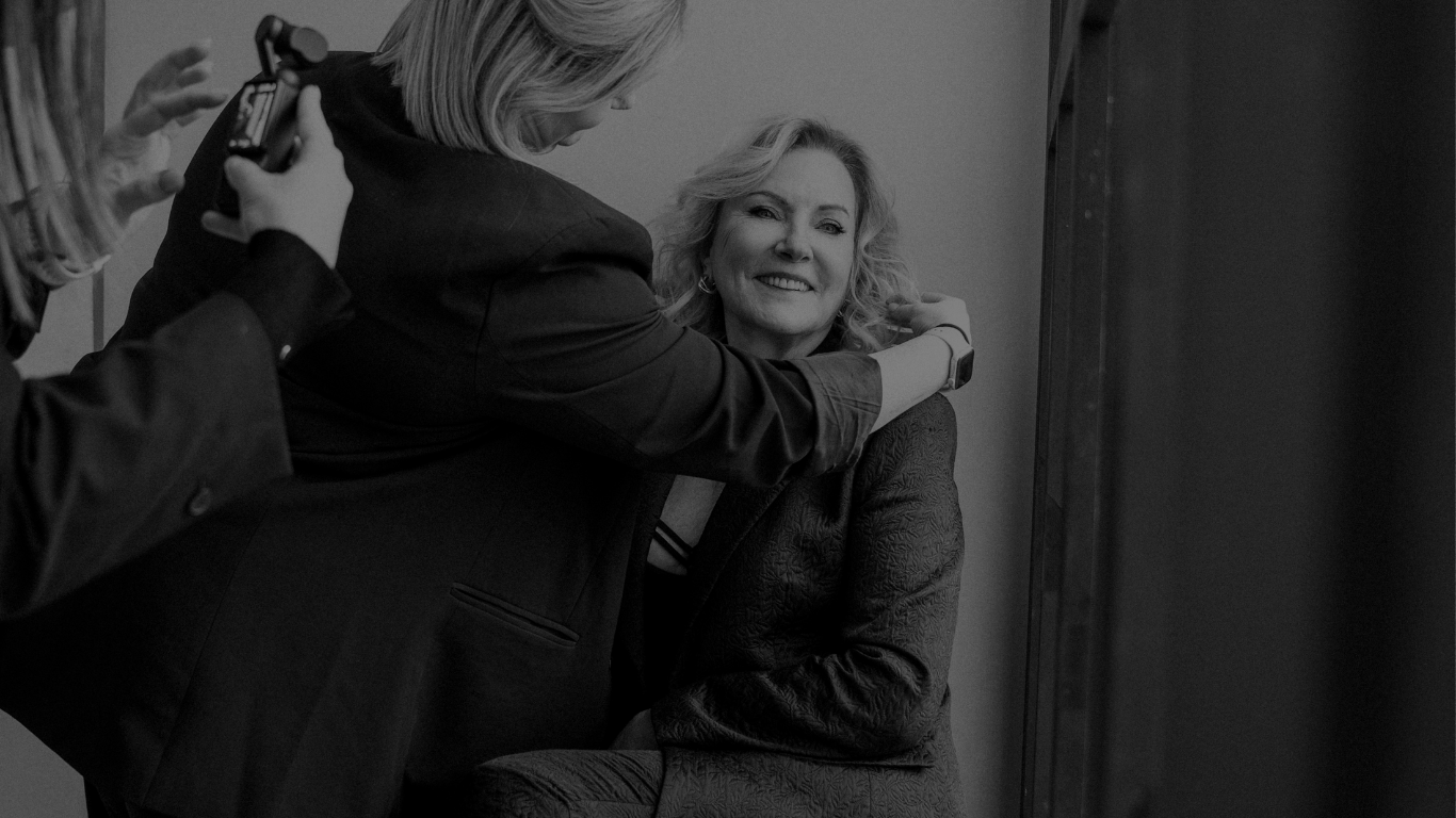 Een vrouw die glimlacht terwijl ze een knuffel krijgt van een andere vrouw in een professionele setting, met een derde persoon die een foto maakt.