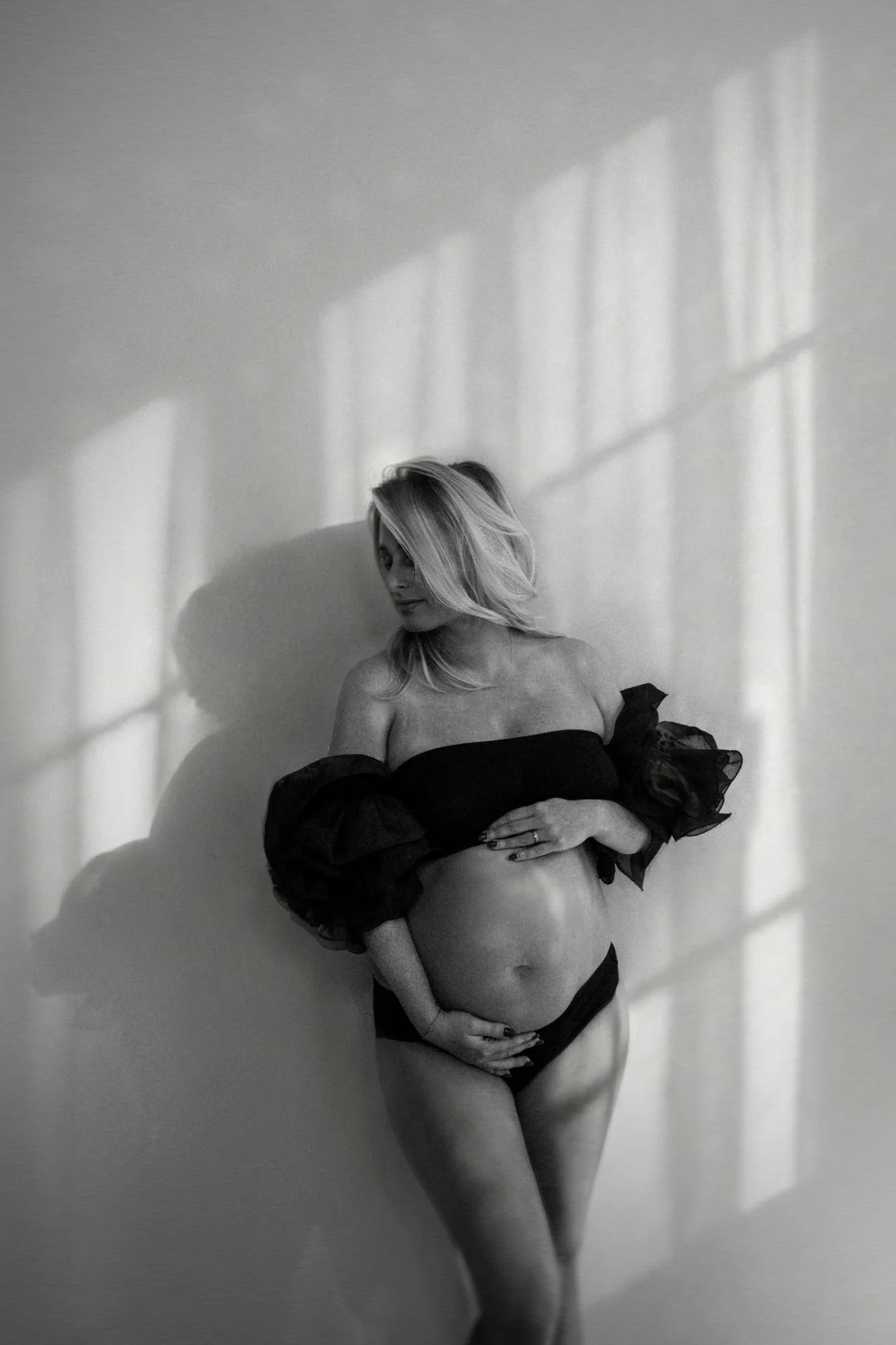 Black-and-white maternity portrait of a pregnant woman in a strapless black dress standing against a sunlit wall.