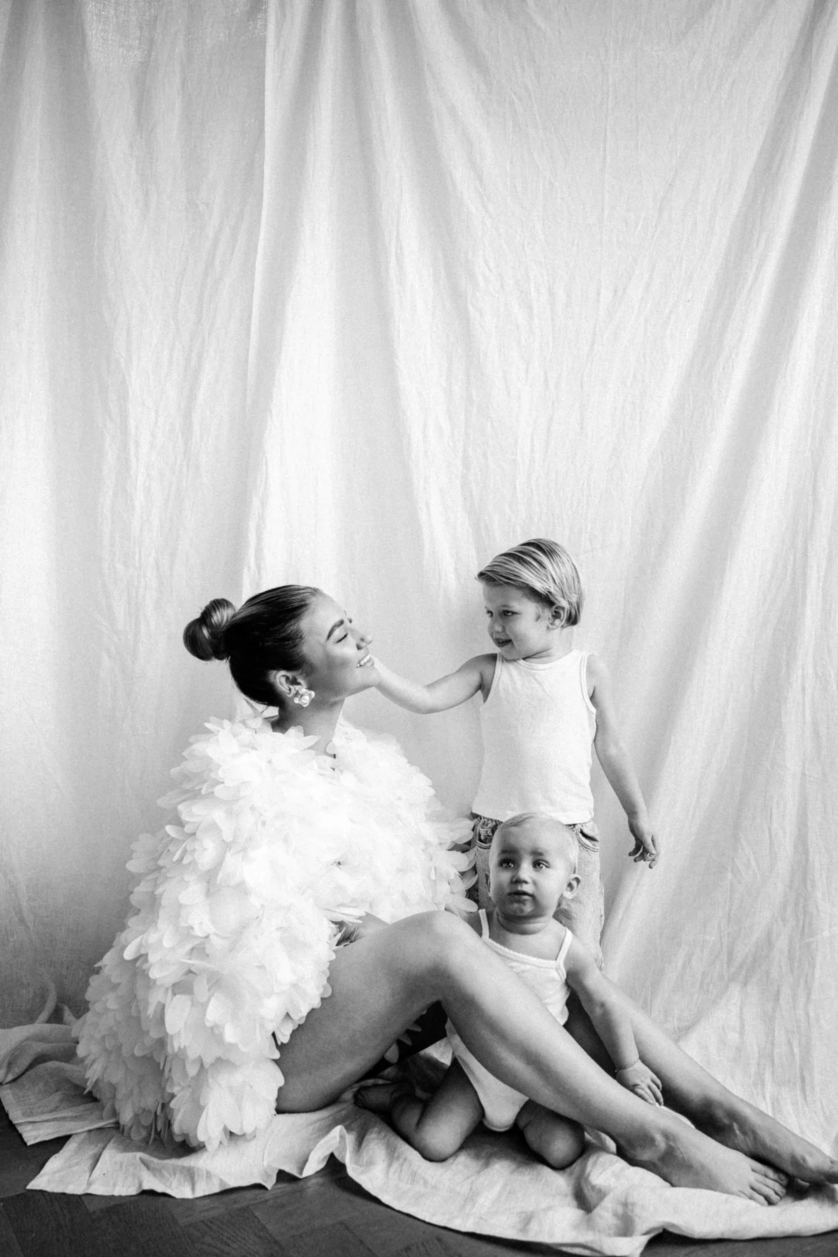 A woman wearing a fluffy white dress with large petal-like fabric pieces sits on the floor, smiling at three young children nearby. One child, a girl, stands touching the woman's face, while a boy and another girl sit close at her feet, all in front of a plain white backdrop.