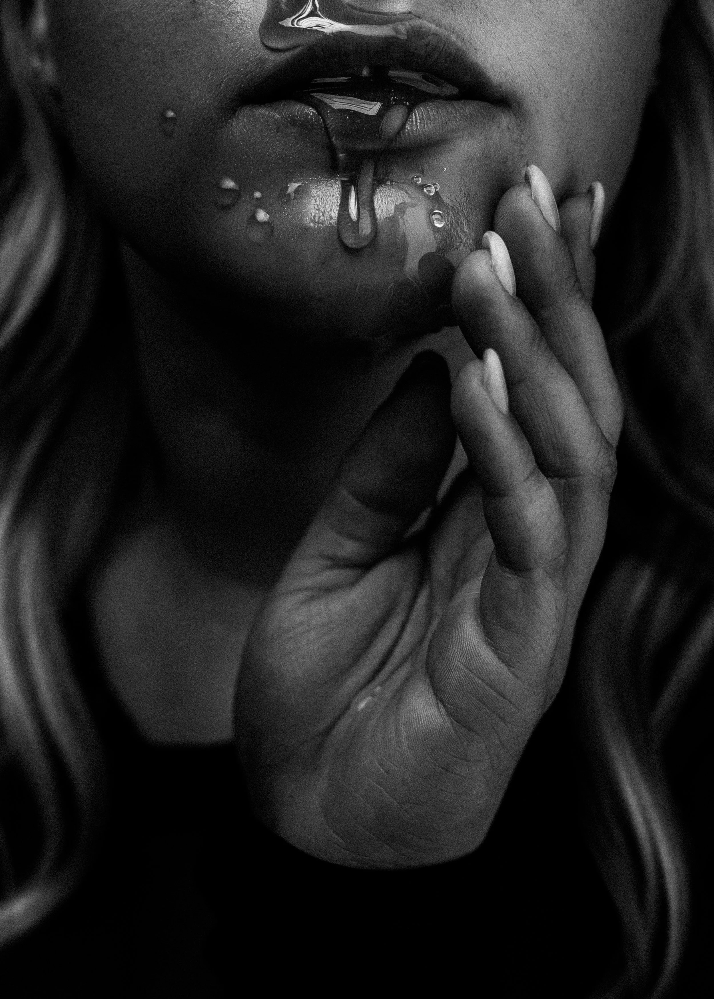 Close-up zwart-wit foto van de lippen en een deel van het gezicht van een vrouw, met haar vingers bij haar mond en een beetje vloeistof die langs haar lippen loopt.