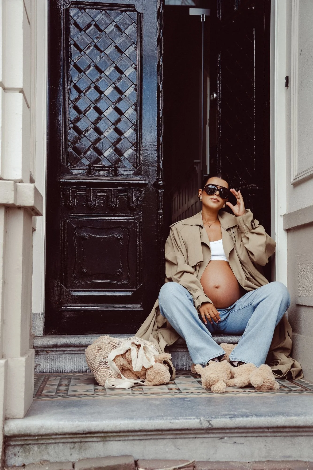 Pregnant woman in sunglasses and a beige trench coat sitting on a doorstep beside a black ornate front door.