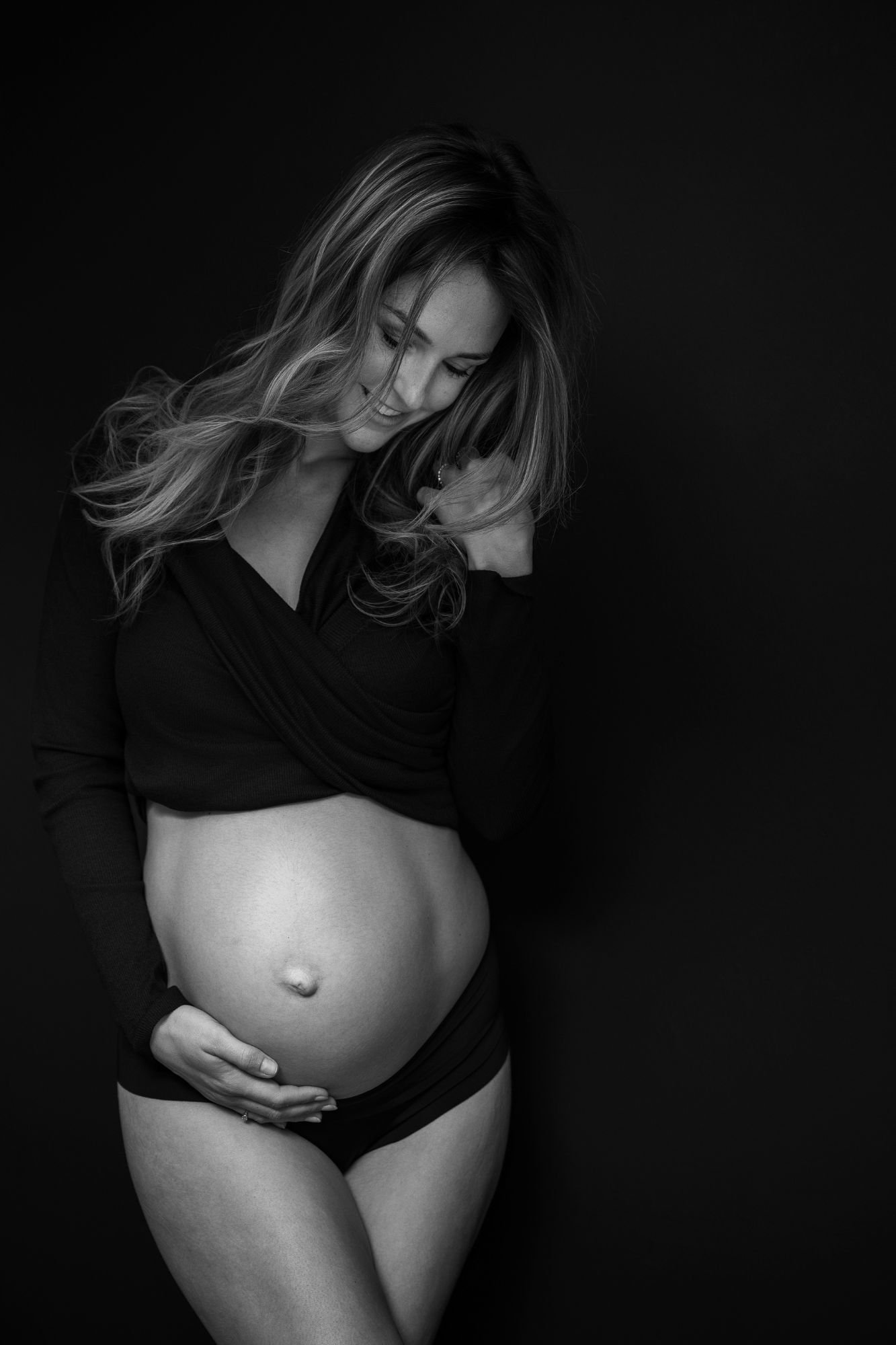 Black-and-white studio portrait of a pregnant woman cradling her belly against a dark background