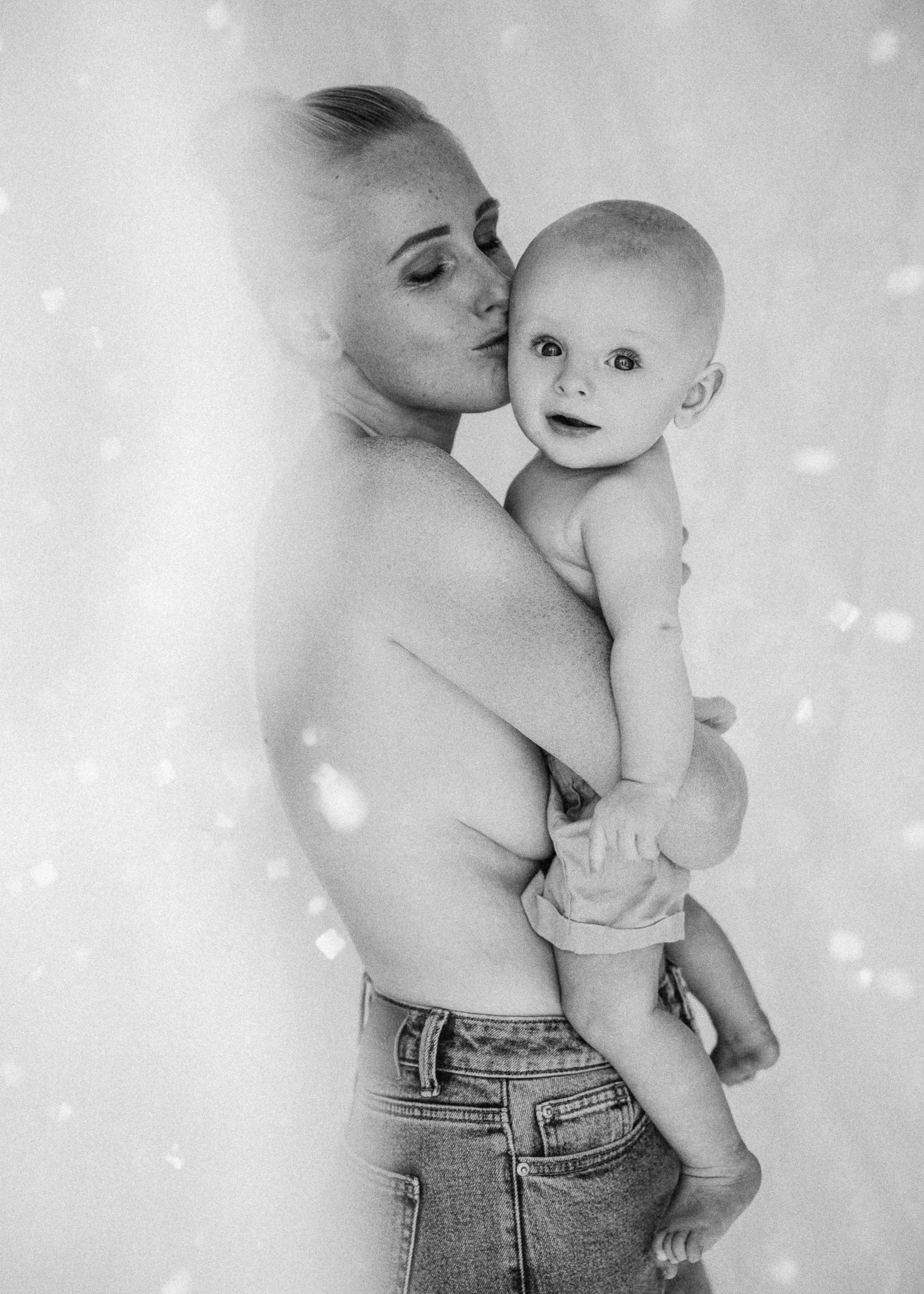 A woman with short hair holding a baby boy with a shaved head. The woman is kissing the baby on the cheek. Both are shirtless, and the woman is wearing jeans. The photo is in black and white with bokeh effects.