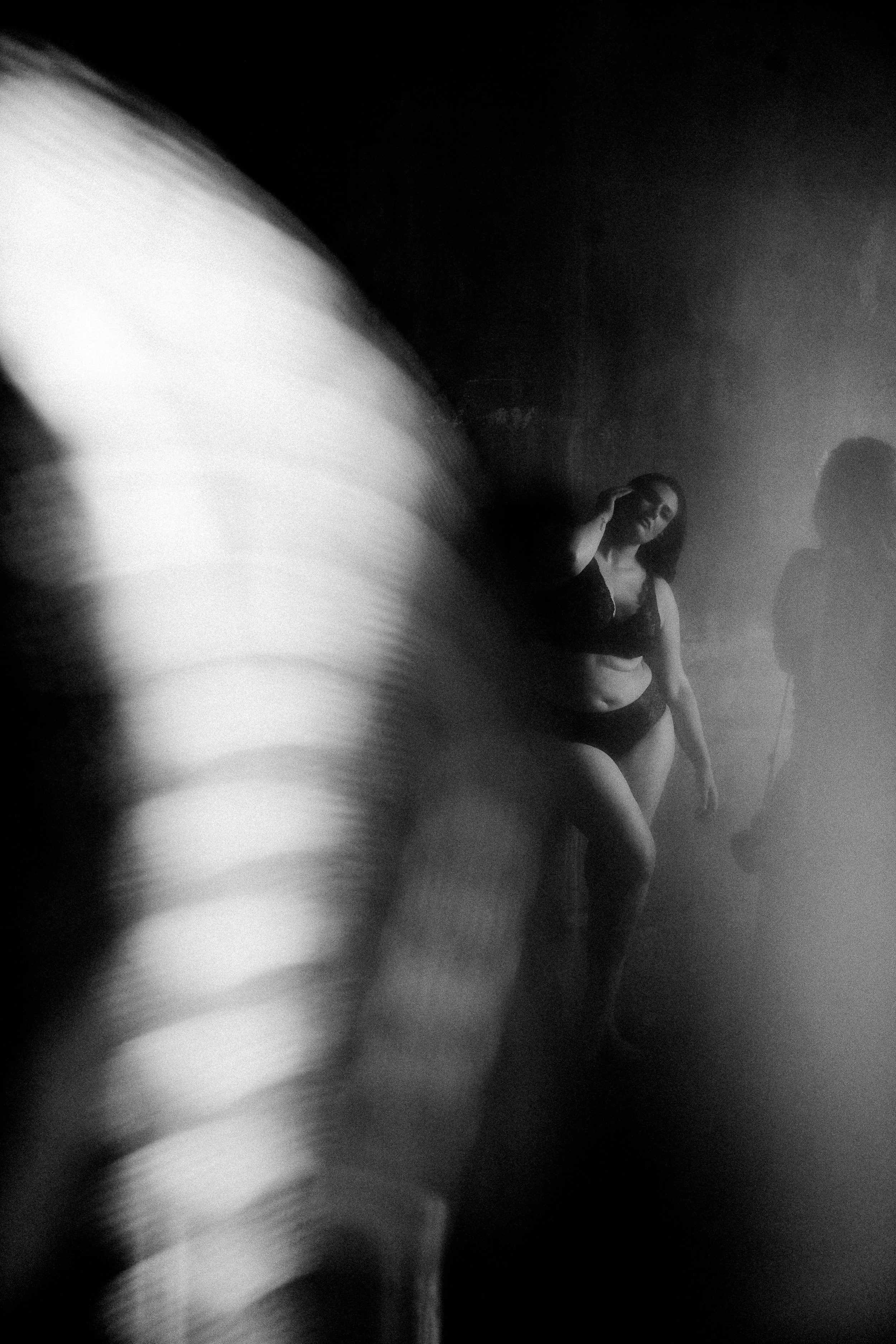 Vrouw in donkere lingerie staand in een nevelige of rookachtige setting, met een vervaagde figuur op de voorgrond en een silhouet op de achtergrond.