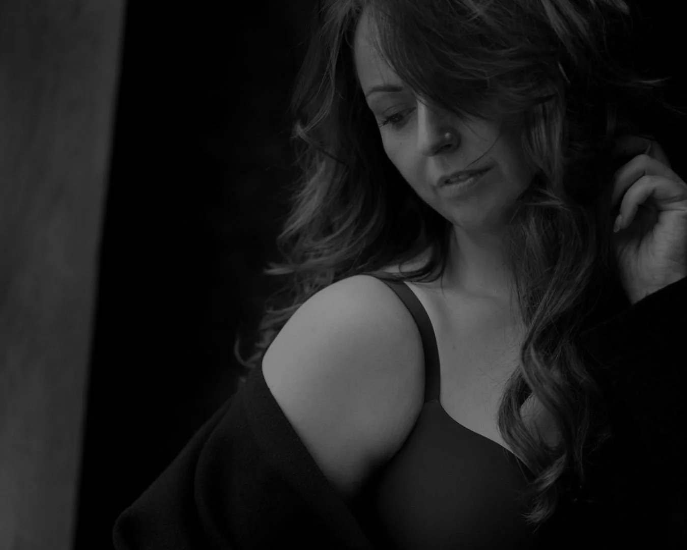 A black and white photo of a woman with wavy hair, looking downward, with her hand near her face, wearing a strap top and jacket.