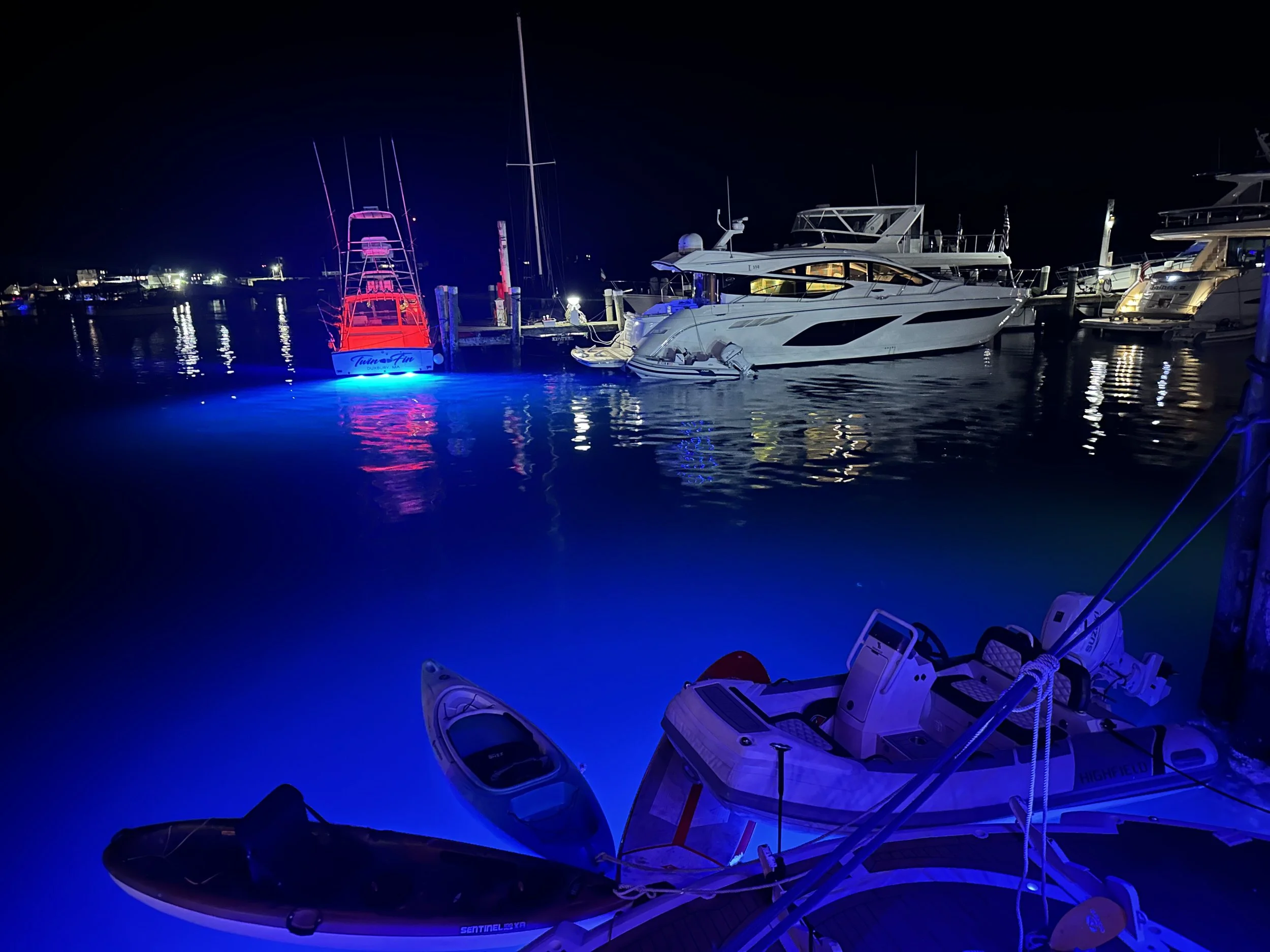 Nighttime scene at a marina with illuminated boats and yachts, reflecting on the calm water, featuring a small inflatable boat and kayaks in the foreground.