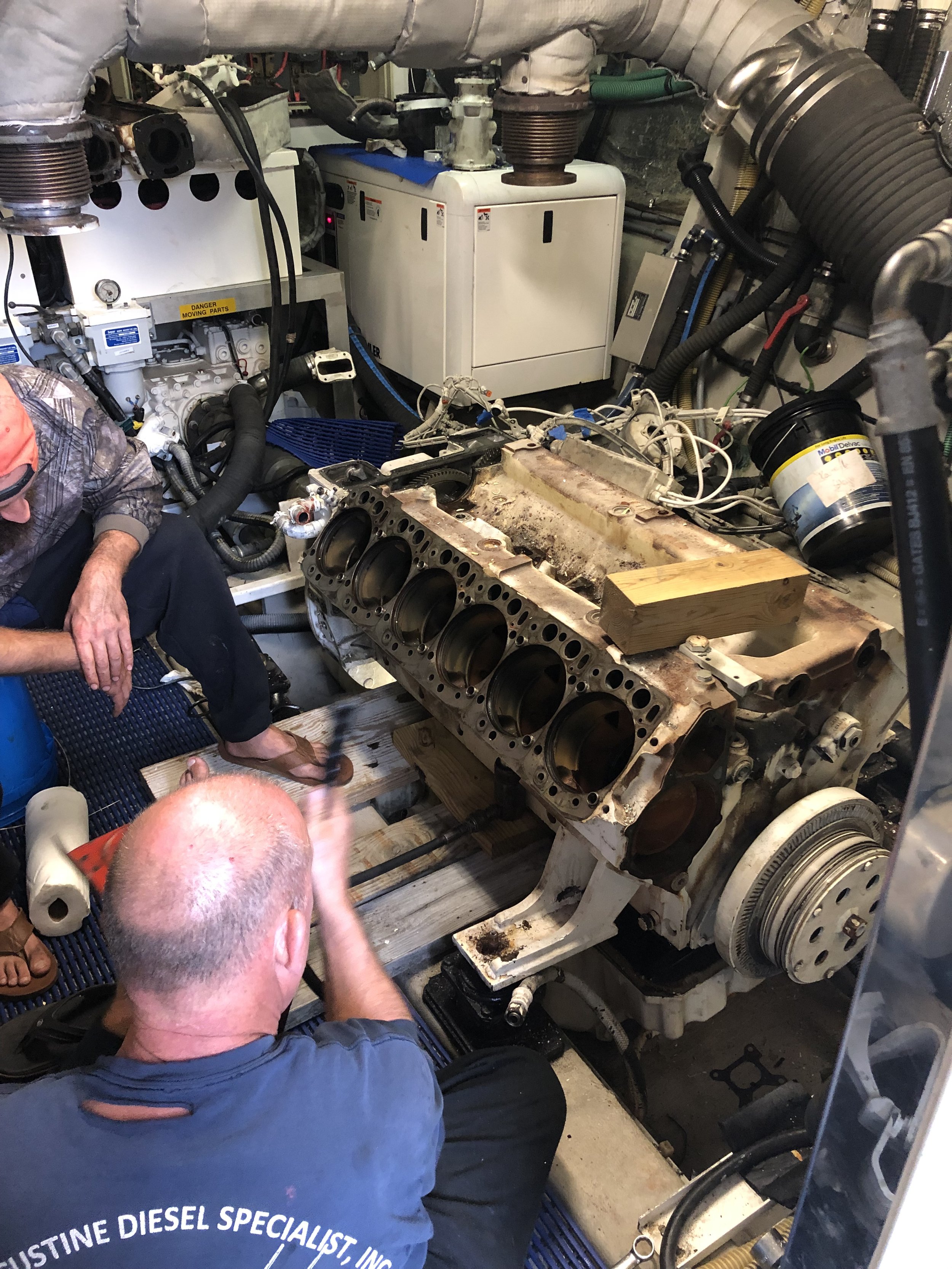 Two mechanics working on a large engine block in a confined workshop space. One mechanic is kneeling, the other sitting on the floor, both focused on the engine, which is clamped onto wooden supports. The engine appears to be partially disassembled with cylinders visible.