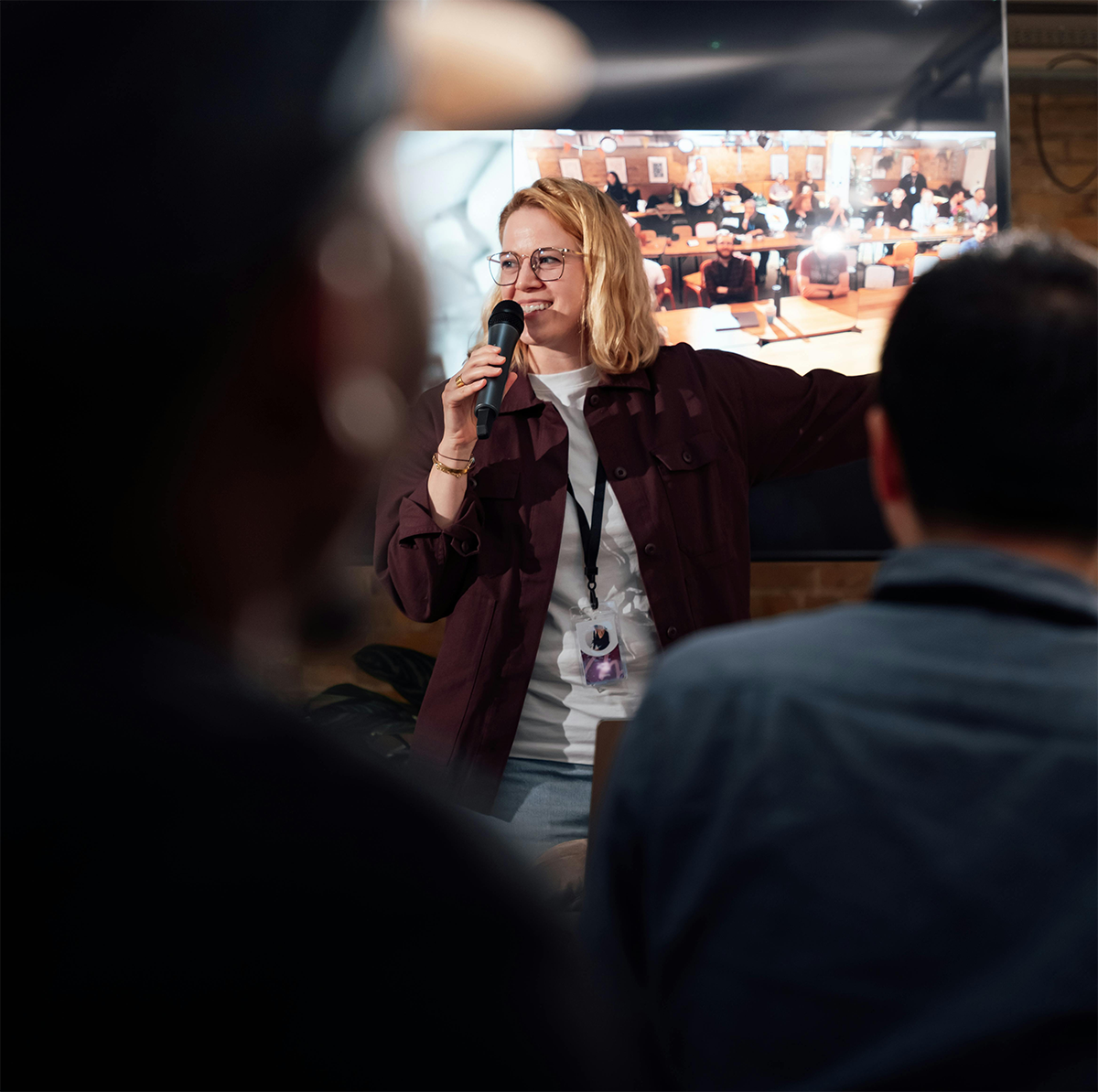 A woman with glasses and blonde hair speaking into a microphone during a presentation or lecture, with a large screen displaying her image in the background, in a room with an audience.