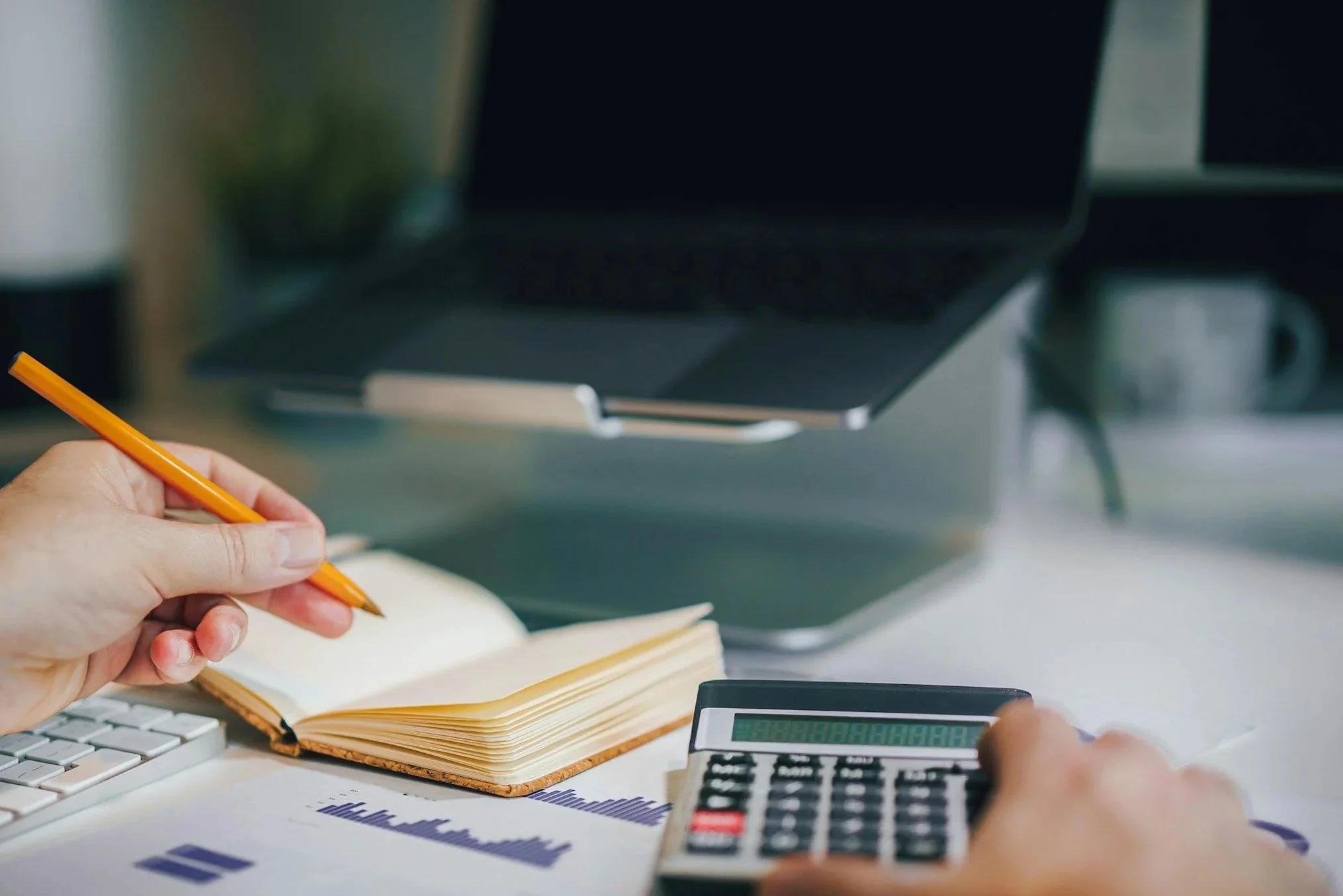 A person working at a desk with a computer, calculator, open notebook, and printed graphs, holding an orange pencil.