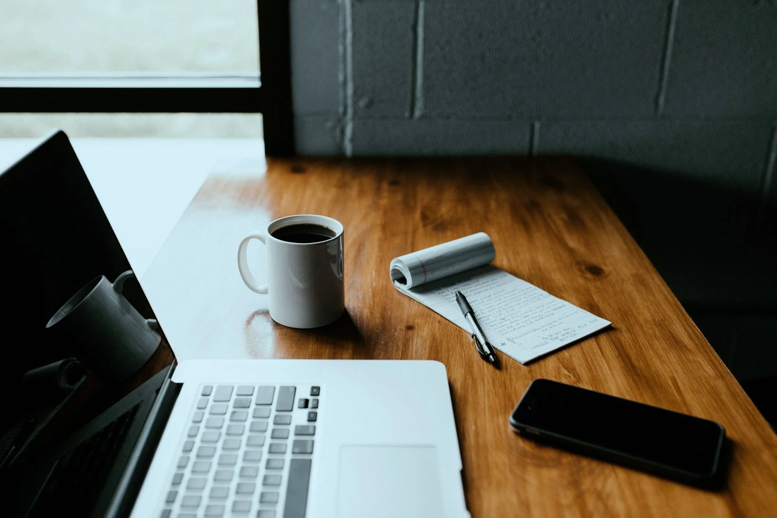 A workspace with a laptop, a coffee mug, a notepad with a pen, and a smartphone on a wooden table near a window.