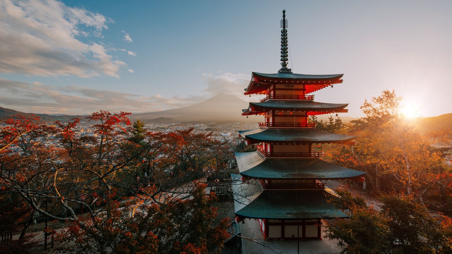 A traditional Japanese five-story pagoda with a backdrop of Mount Fuji, autumn trees, and the setting sun.