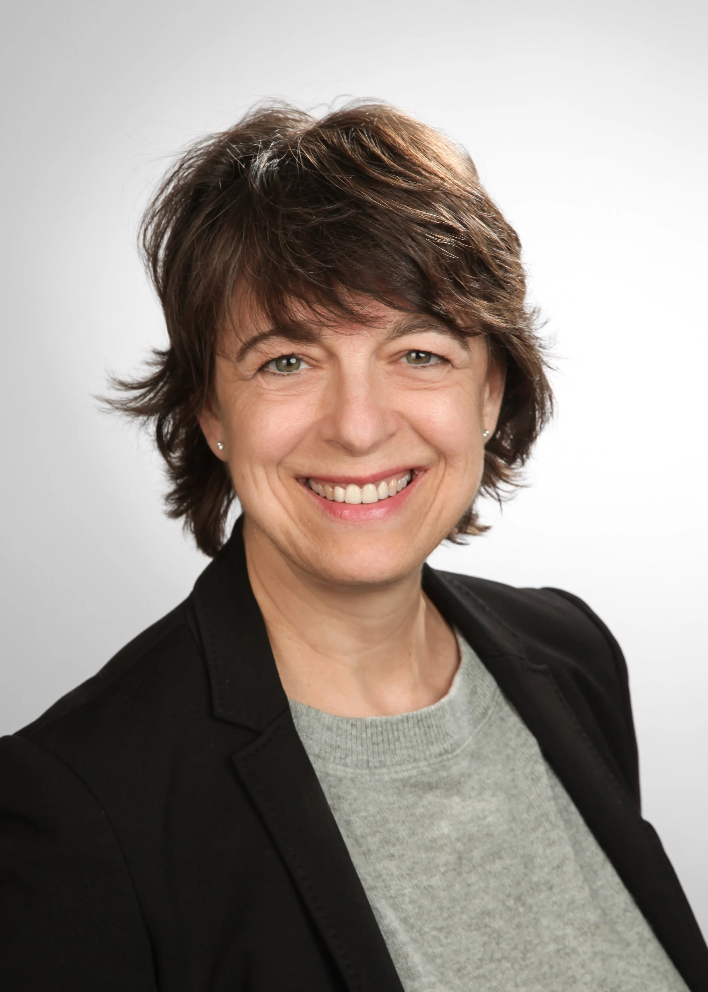 eine lachende Frau mit braunem, lockigem Haar in einem schwarzen Blazer und grauem Hemd