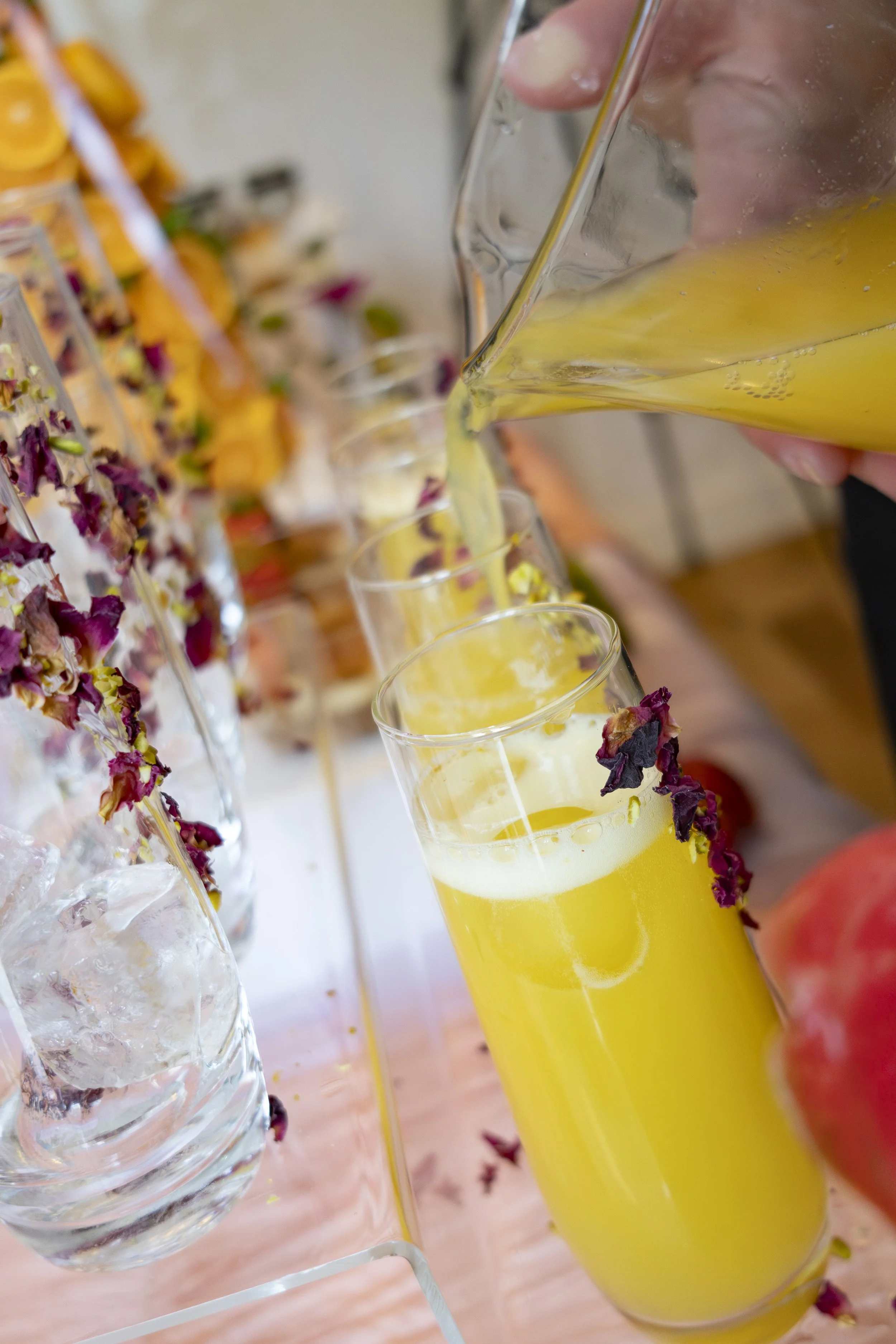 A hand pouring yellow beverage into a glass, with other glasses filled with similar drinks on a table decorated with flower petals and ice.