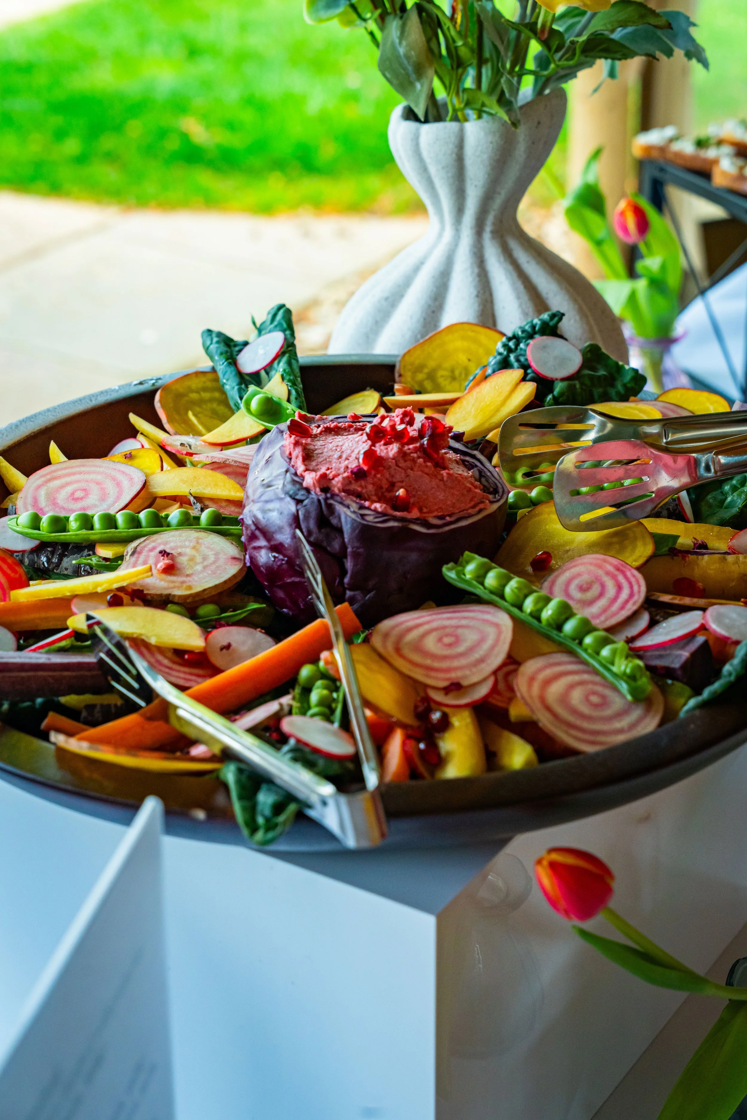 Colorful vegetable platter with sliced radishes, pickles, carrots, and leafy greens, positioned on a table with a decorative vase in the background.