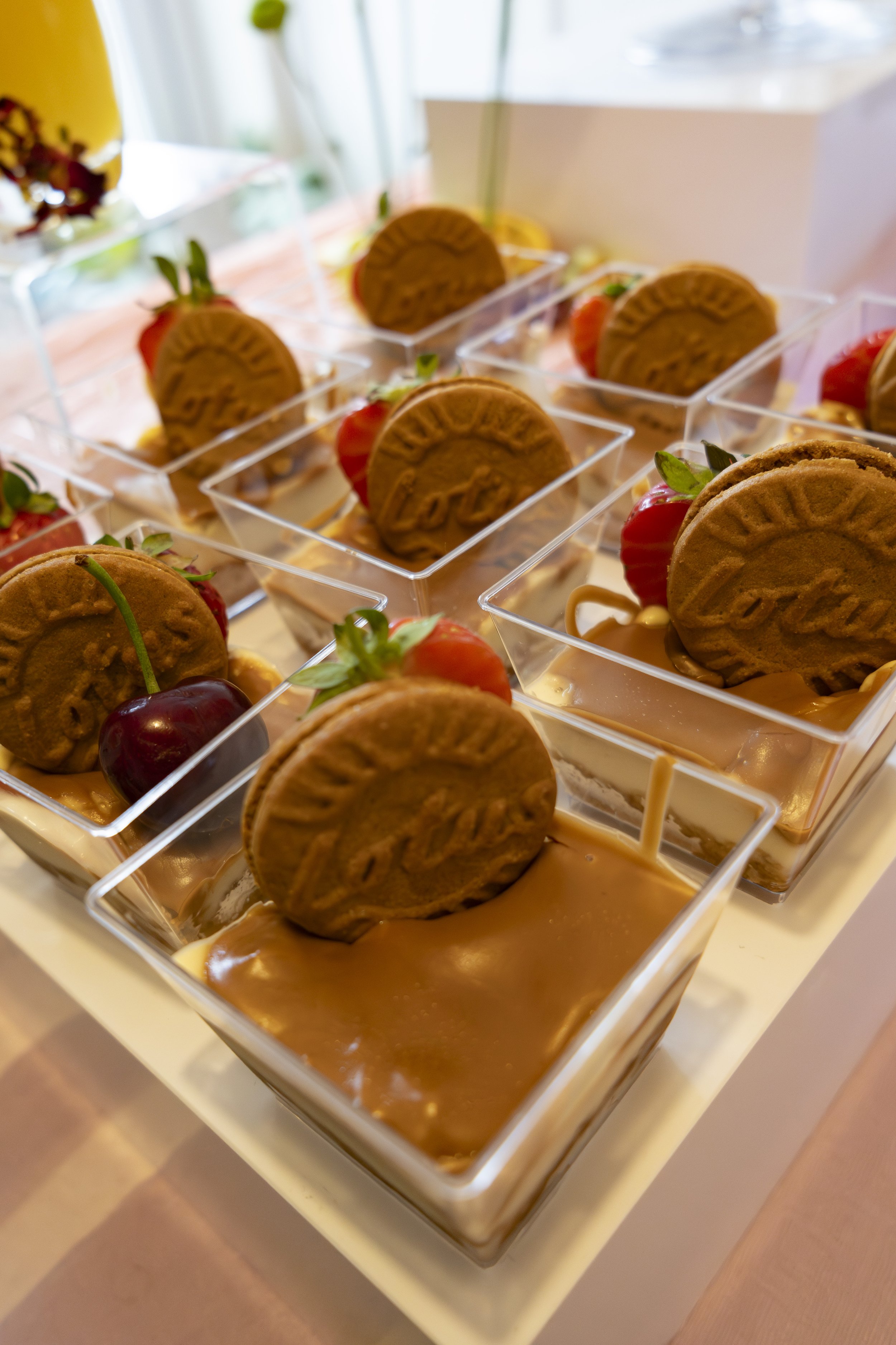 Several glass cups filled with caramel pudding, topped with cookies and strawberries, arranged on a white tray.