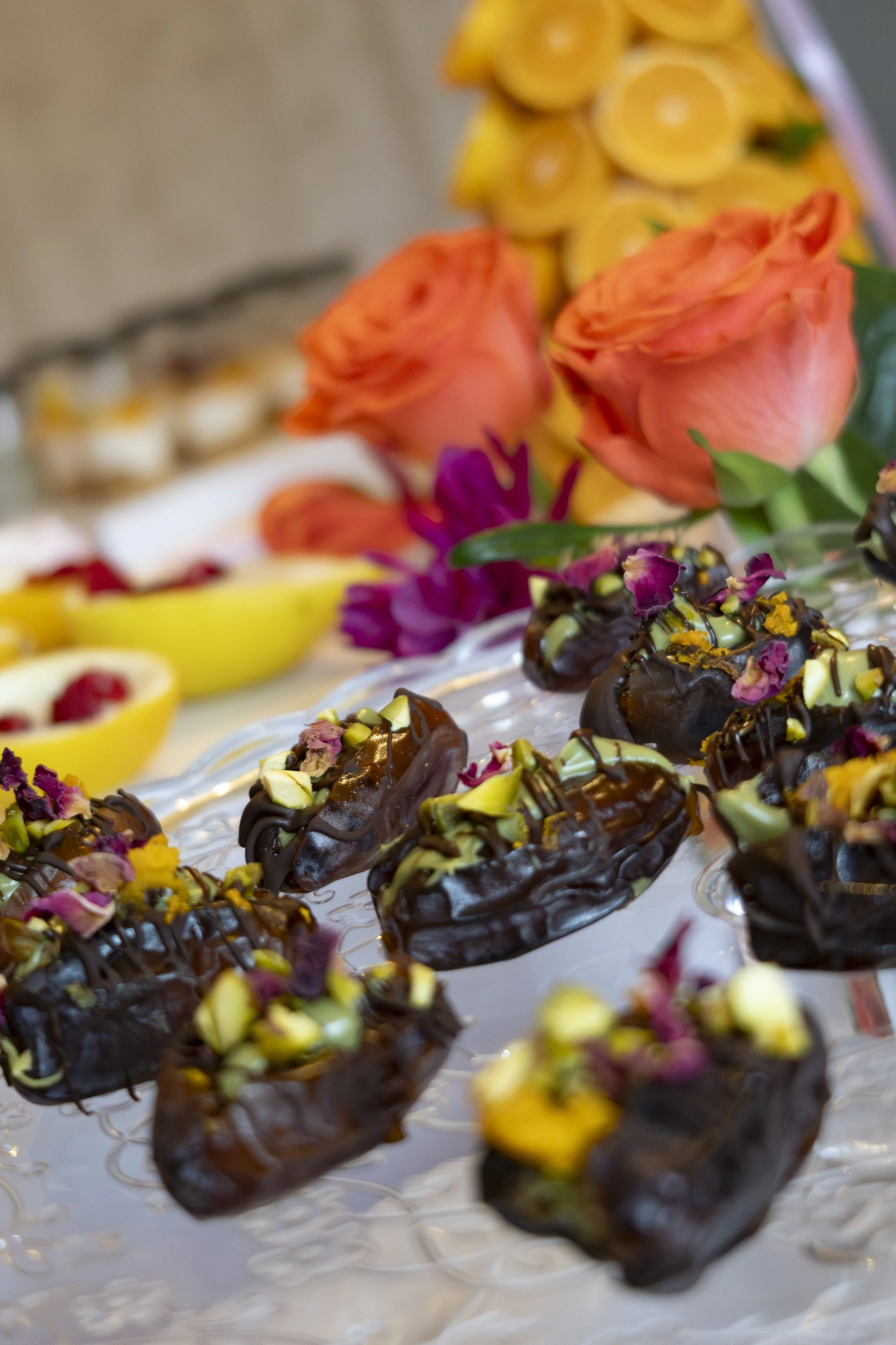 A tray of chocolate éclairs topped with chopped nuts and pink flower petals, with orange roses and yellow flowers in the background.