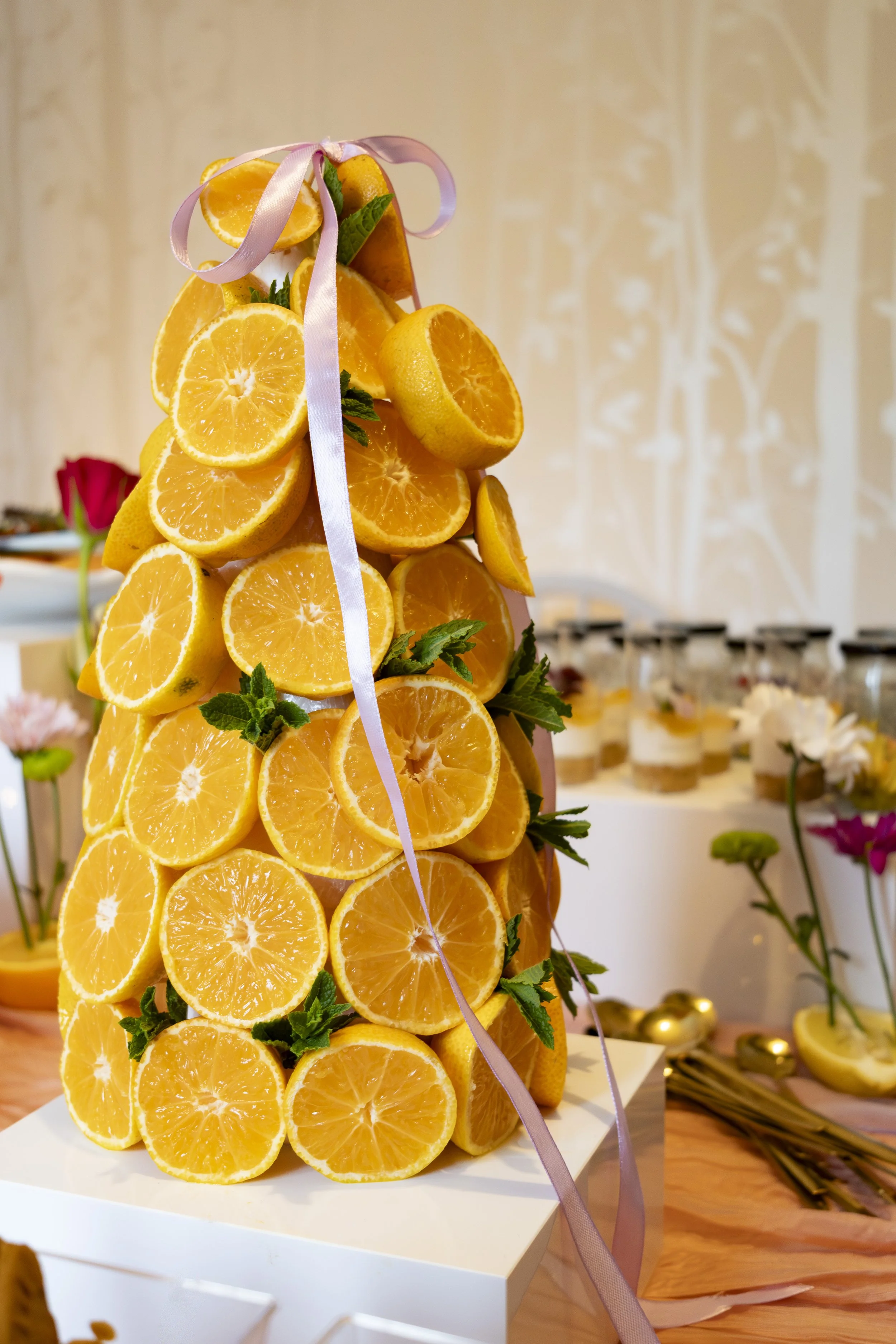 A pyramid-shaped citrus fruit display made of sliced oranges, decorated with green mint leaves and pink ribbon, on a white square base at a celebration event.