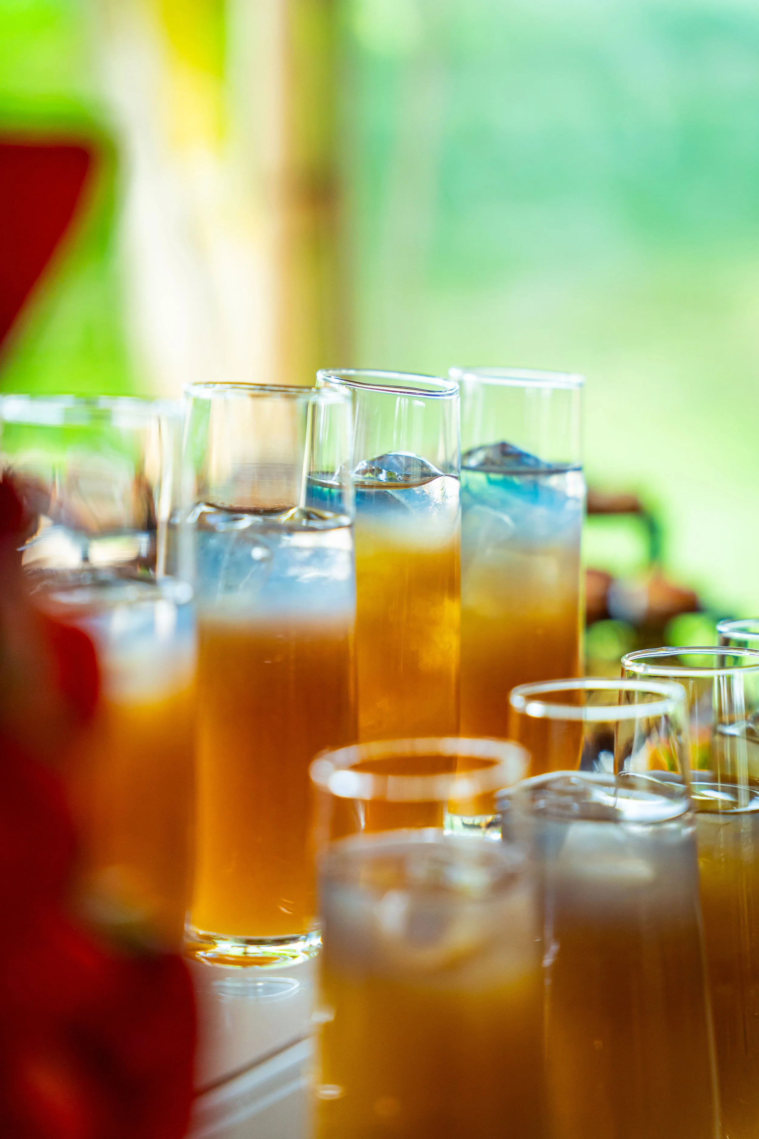 Close-up image of colorful drinks with ice in tall glasses on a table, with a blurred vibrant green background.