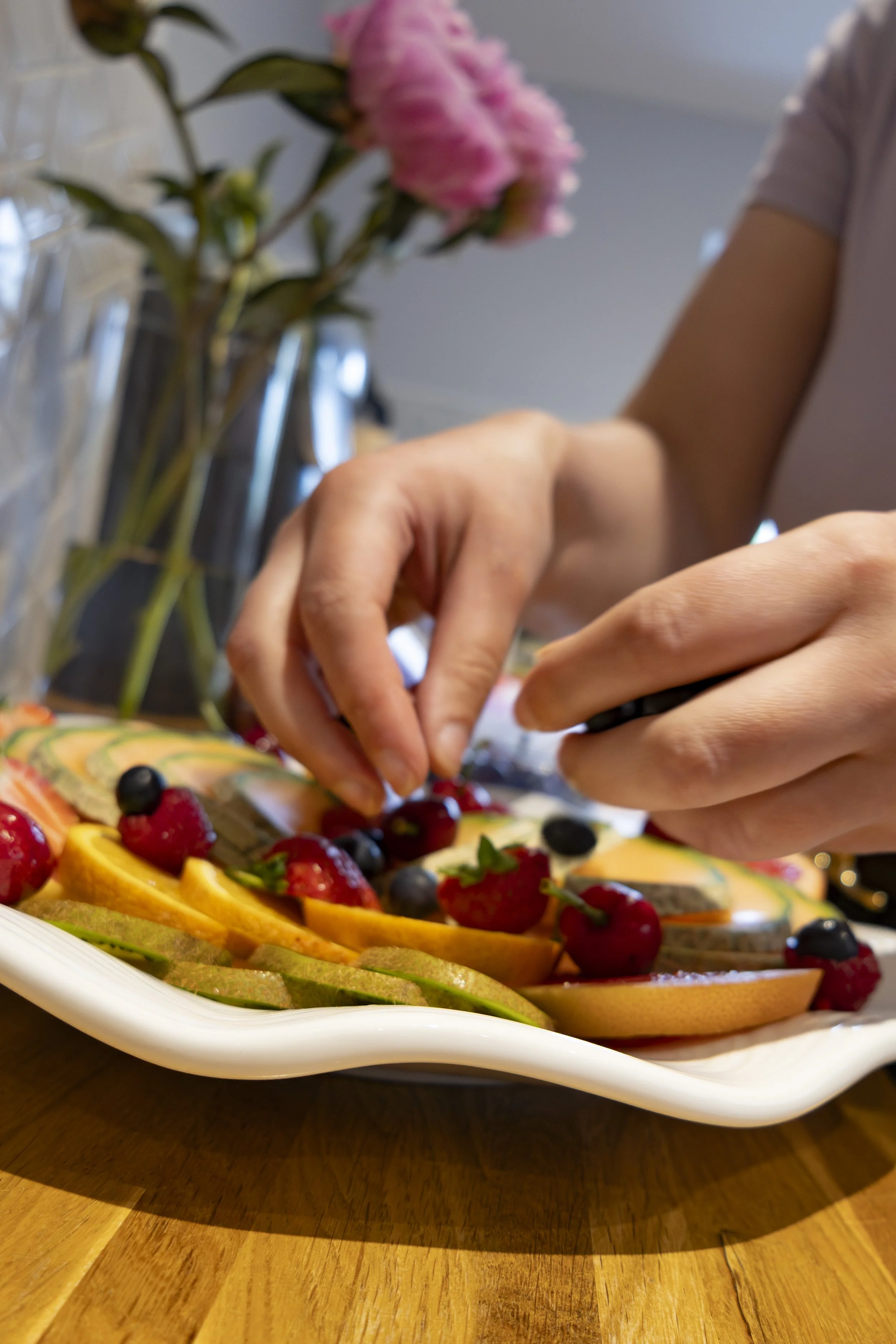 Close-up of hands placing berries on a fruit platter with sliced melon, strawberries, blueberries, and raspberries, on a wooden table.
