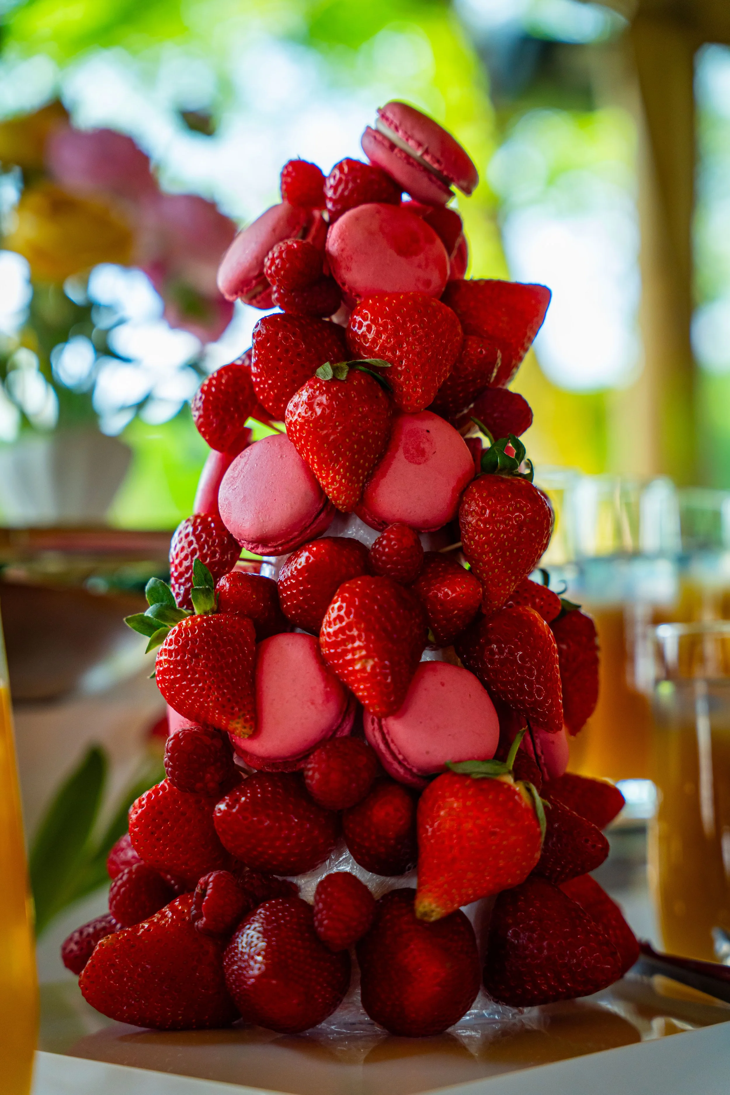 A tall tiered display of pink macarons and fresh strawberries arranged in a cone shape, with a blurred background of green and yellow outdoor scenery.