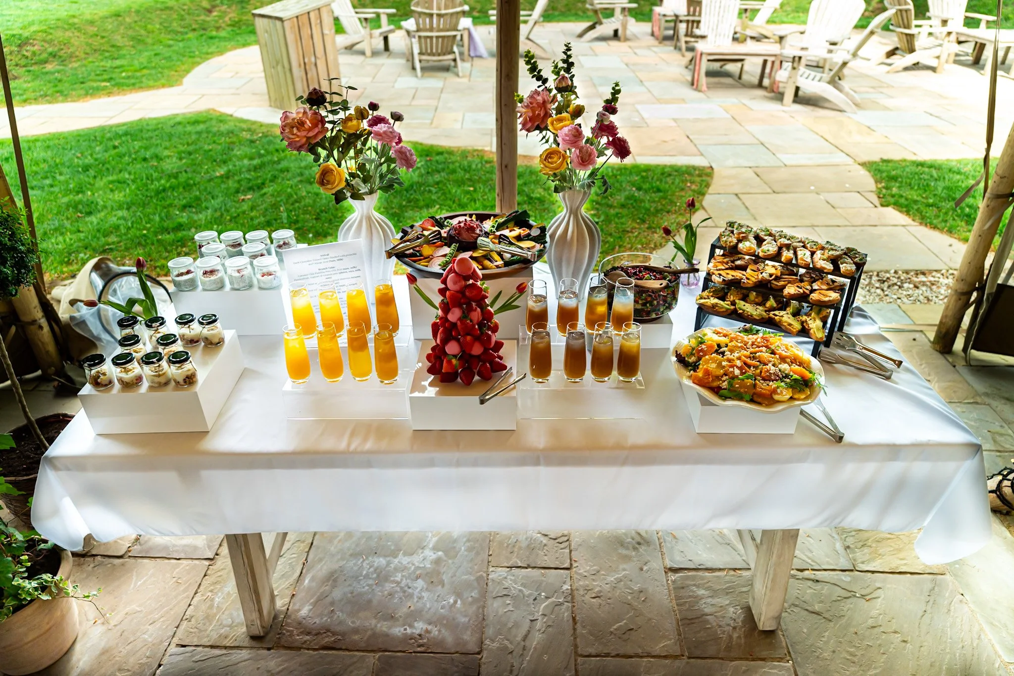 A table set with various desserts, drinks, and floral arrangements, located outdoors on a stone patio with additional chairs in the background.