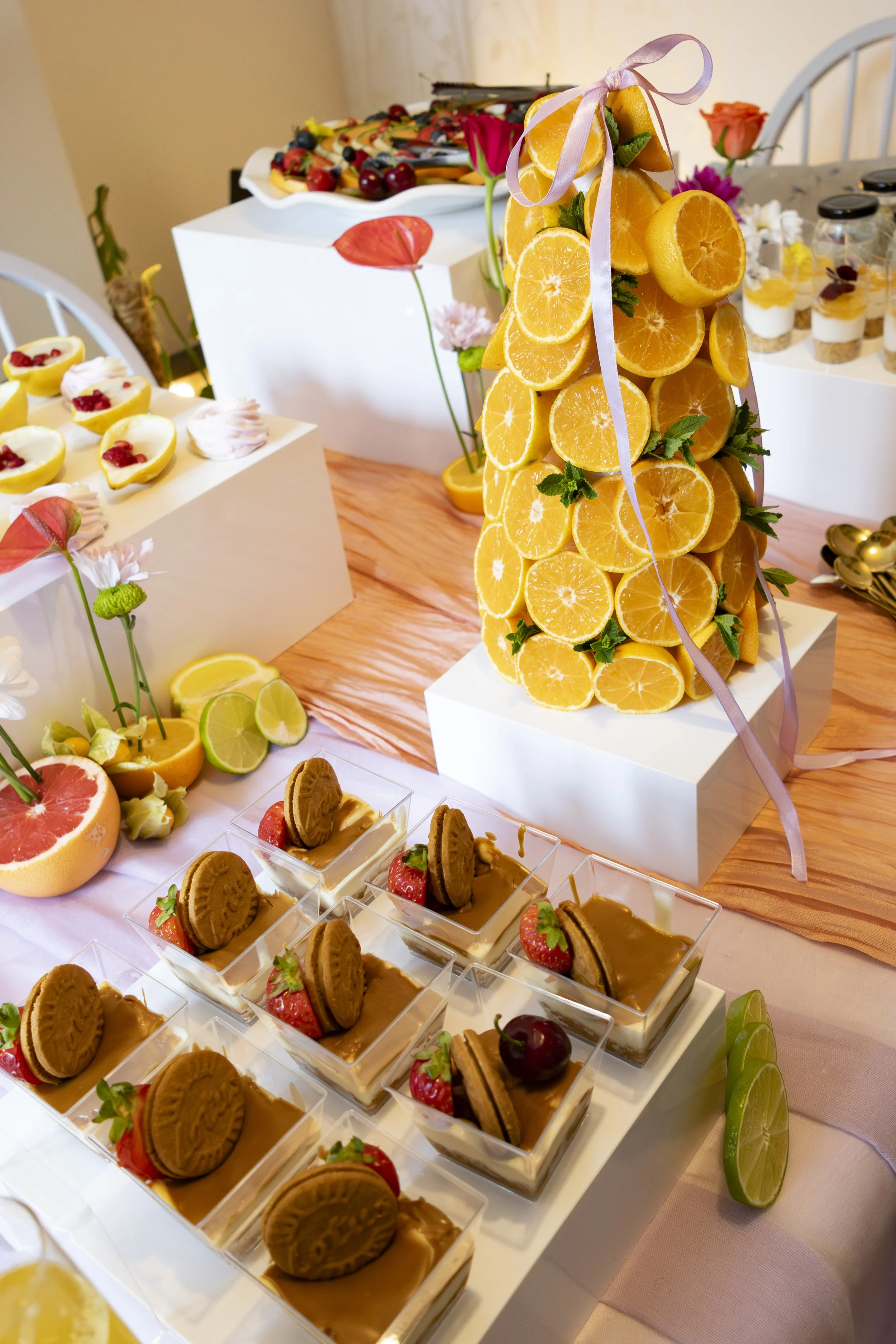 Arrangement of mini desserts including layered pudding cups topped with cookies, strawberries, and chocolate, with slices of oranges, lemons, limes, and garnished with flowers and decorative elements on a table.