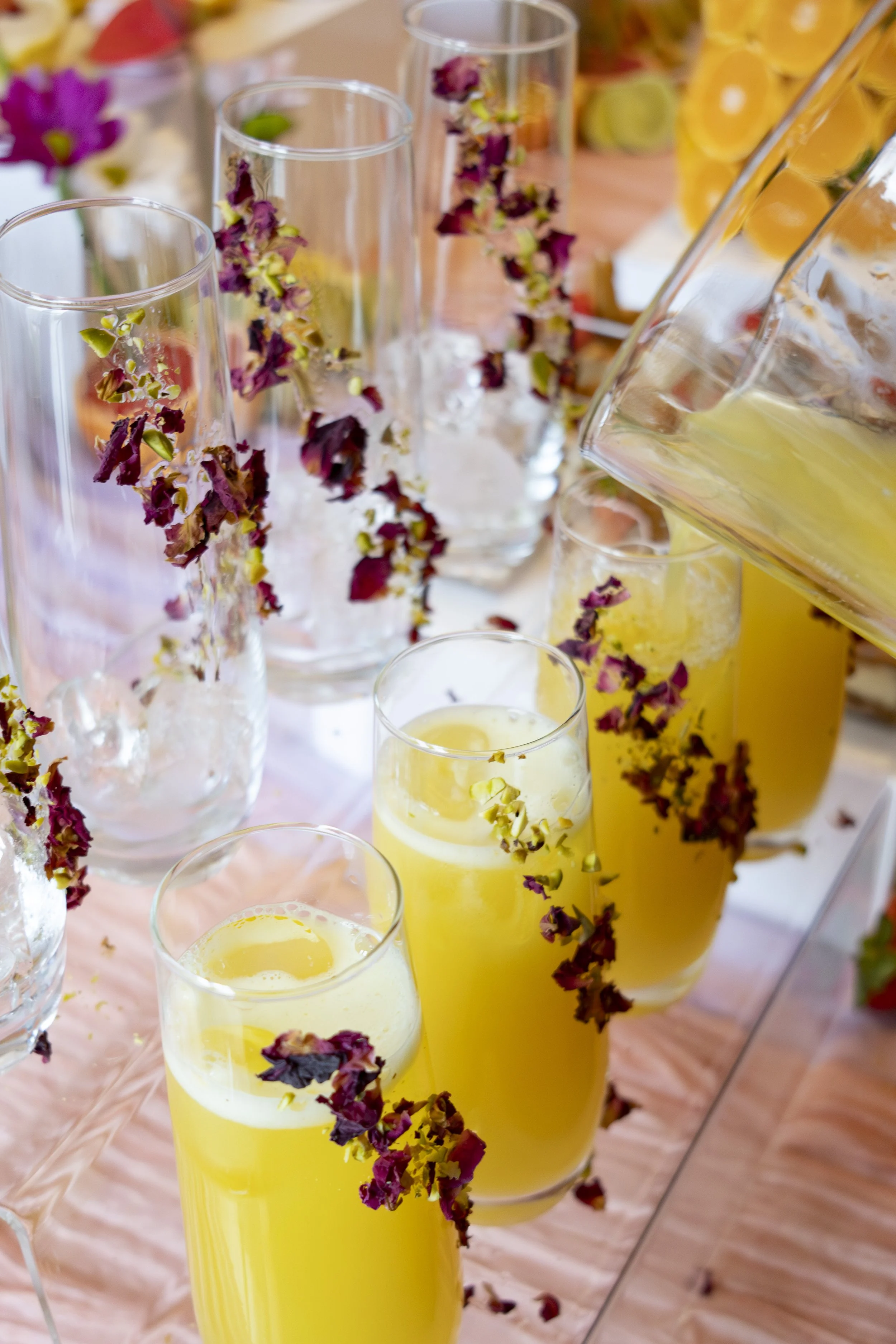 Several tall glasses filled with a yellow beverage, garnished with dried purple flowers, on a table decorated with a pink tablecloth, with a pitcher pouring more of the yellow drink.