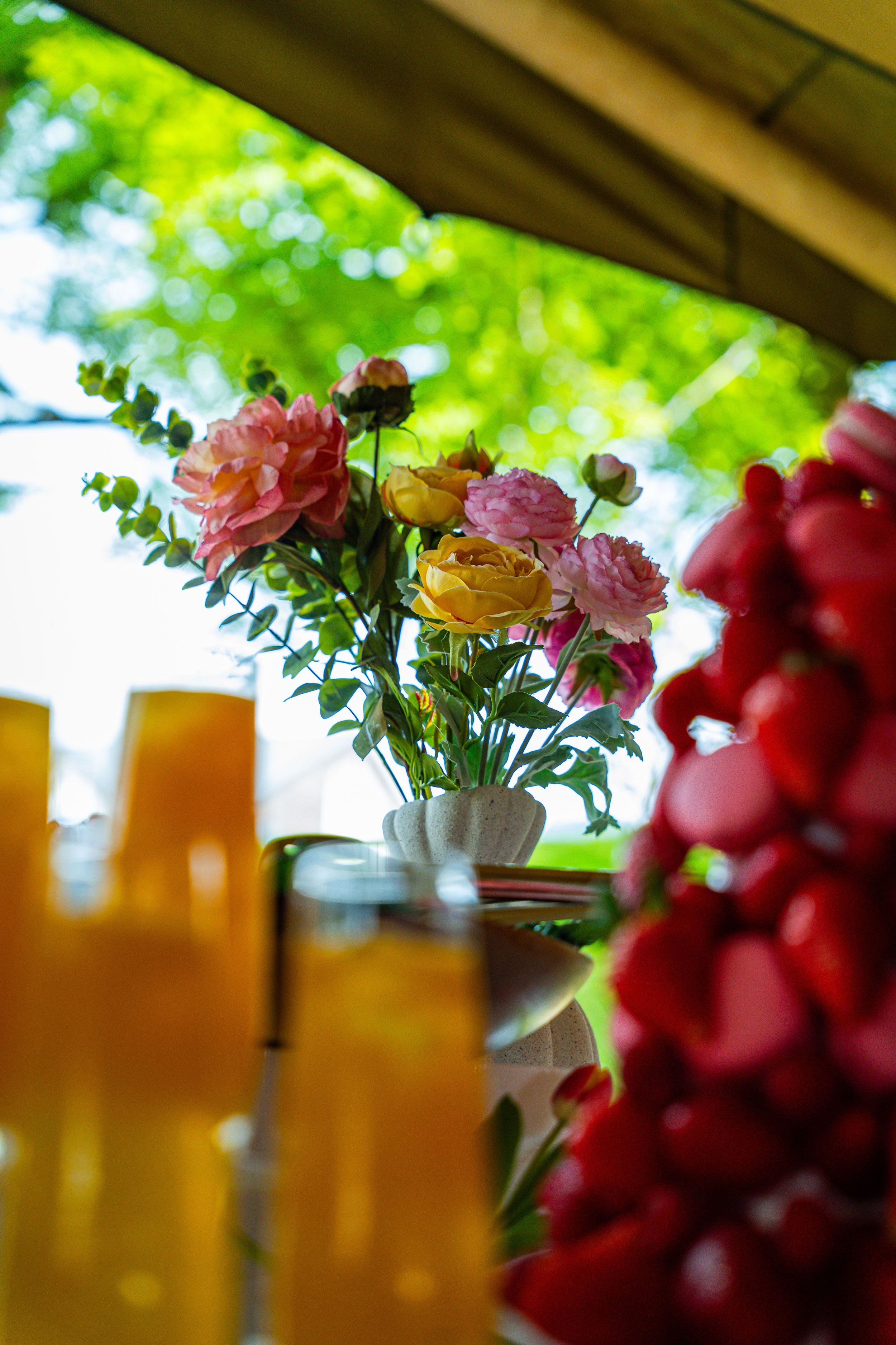 A colorful flower arrangement in a white vase, with pink and yellow flowers, on a table near a window with a green outdoor background. Some yellow objects are visible in the foreground.
