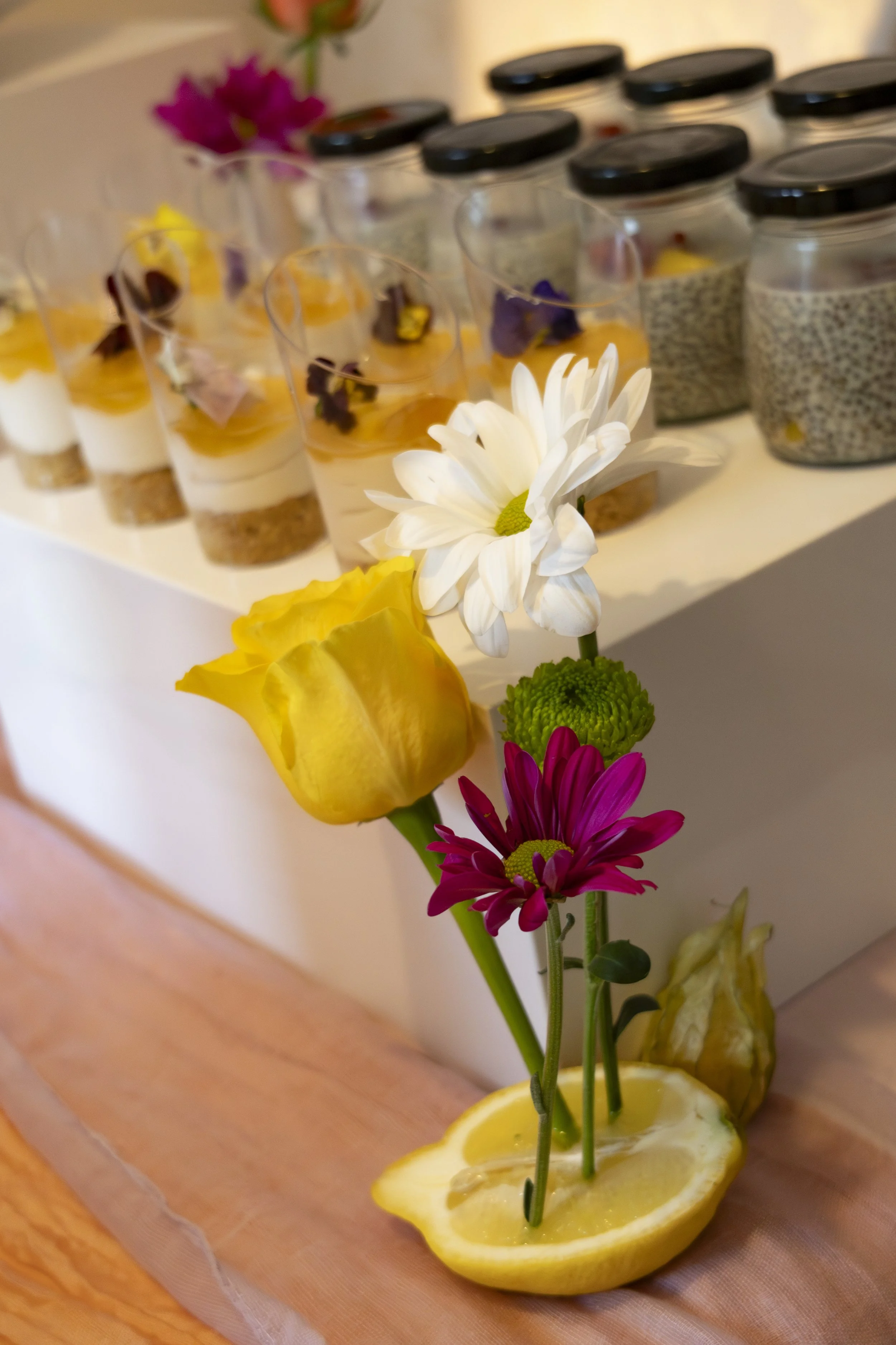 A close-up of decorated desserts in glasses with flowers and lemon slices in the foreground.