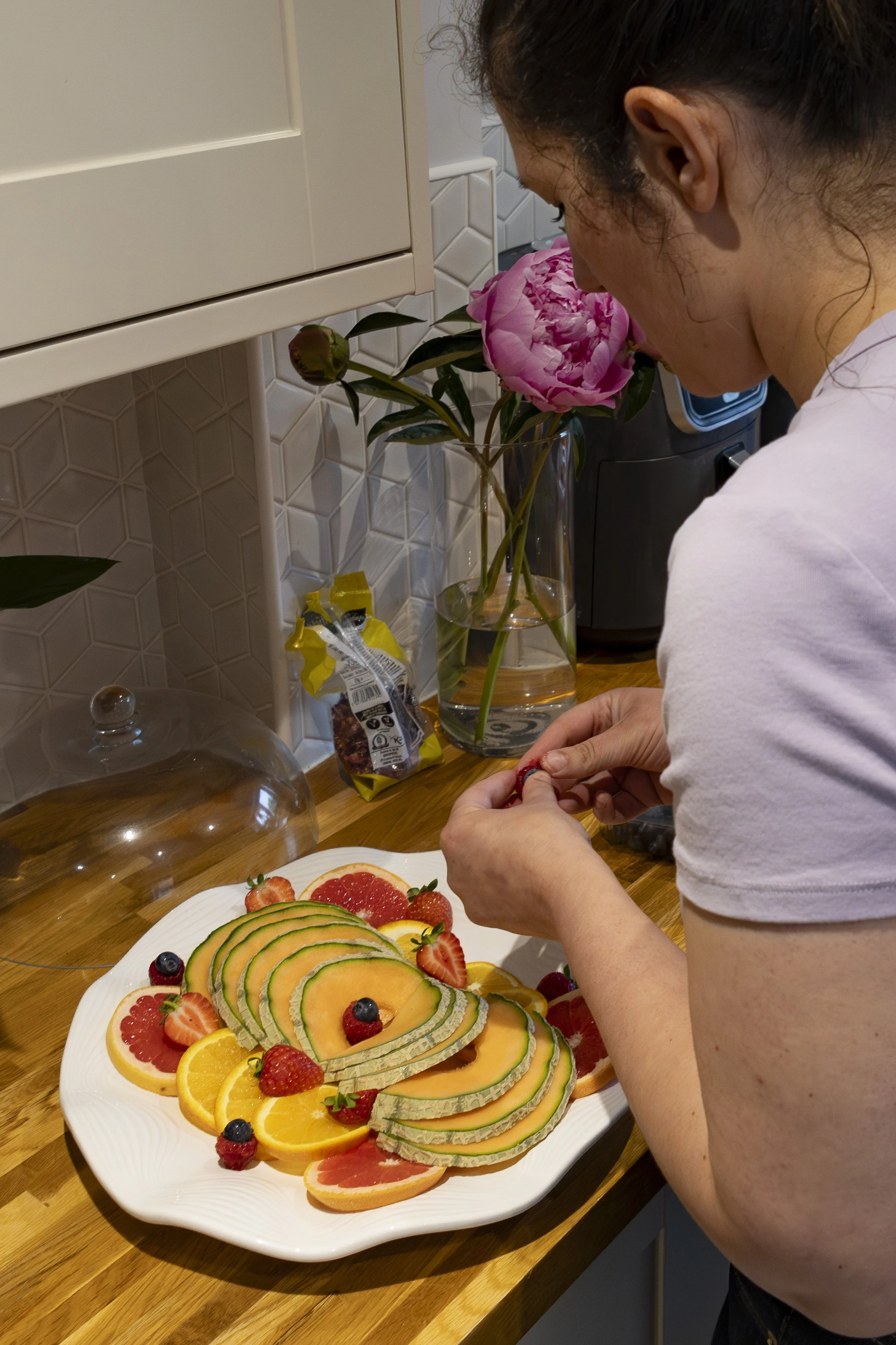 A woman is preparing a fruit platter with sliced melon, grapefruits, oranges, berries, and a decorative flower arrangement on a white plate on a wooden countertop.