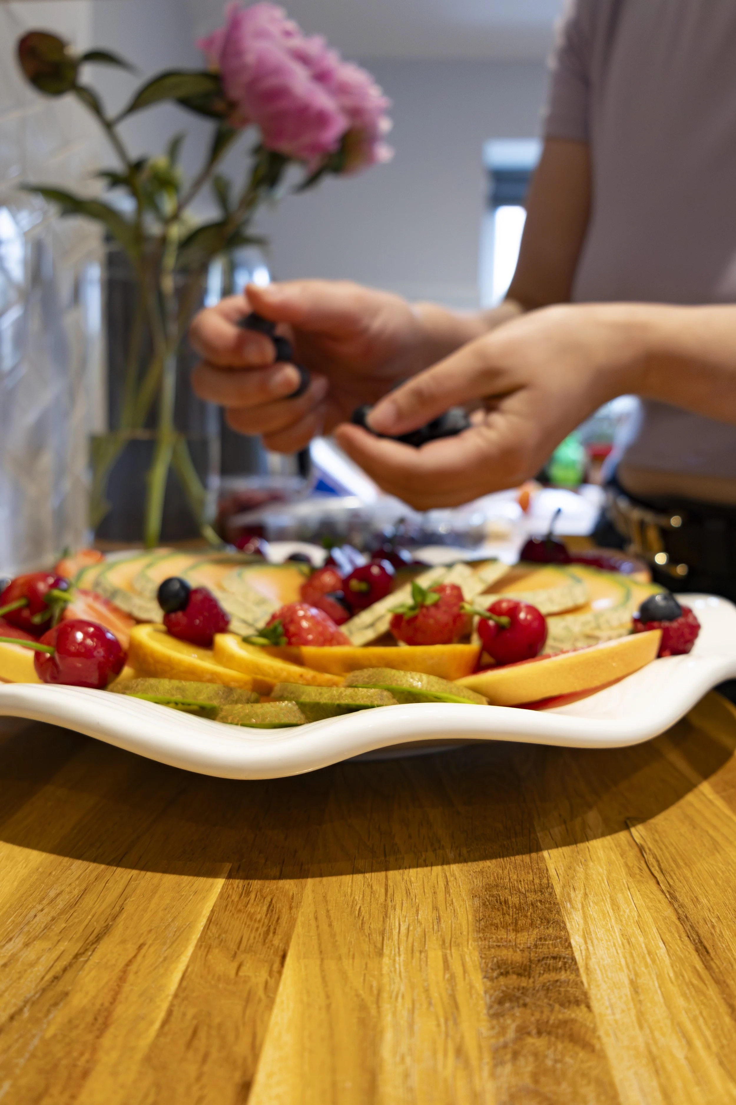 Close-up of a fruit pizza on a white plate, topped with strawberries, blueberries, raspberries, and slices of kiwi, with a person in the background preparing it.