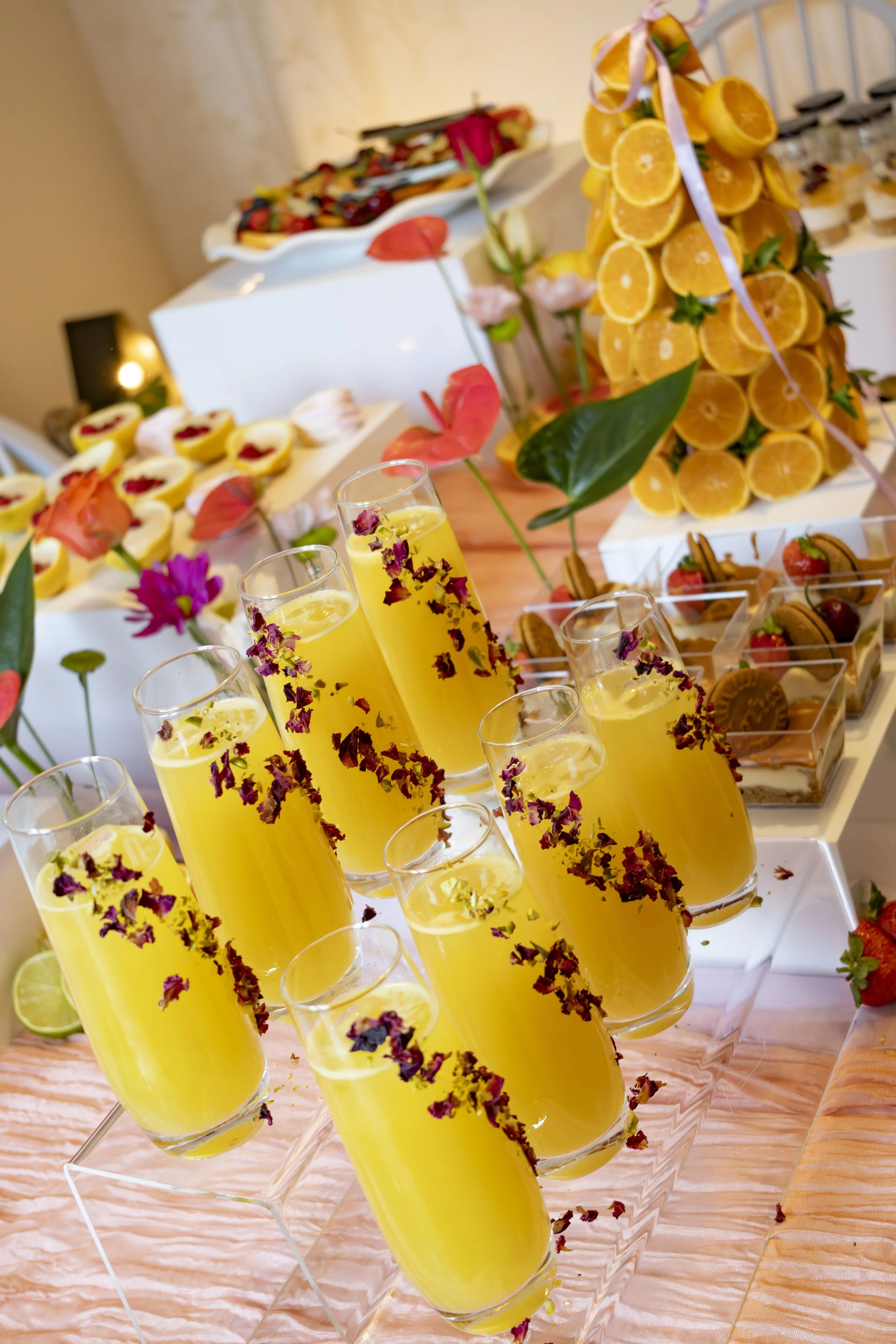 A dessert table featuring tall glasses of yellow drink garnished with flower petals, topped with a large pineapple floral arrangement, with various other desserts and fruit arrangements in the background.