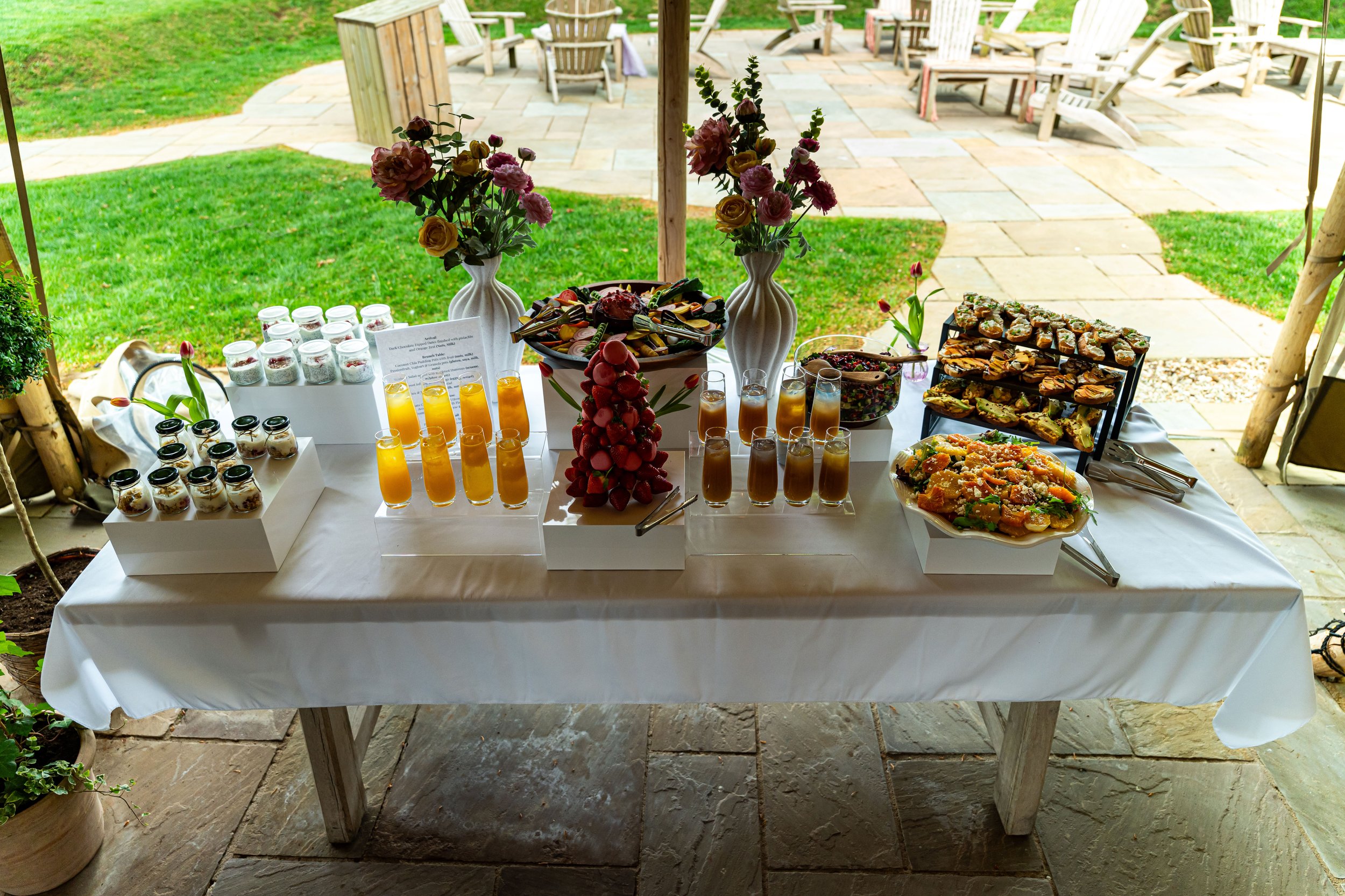 A dessert buffet table with flower arrangements and assorted sweet treats, set outdoors on a patio with lawn and chairs in the background.