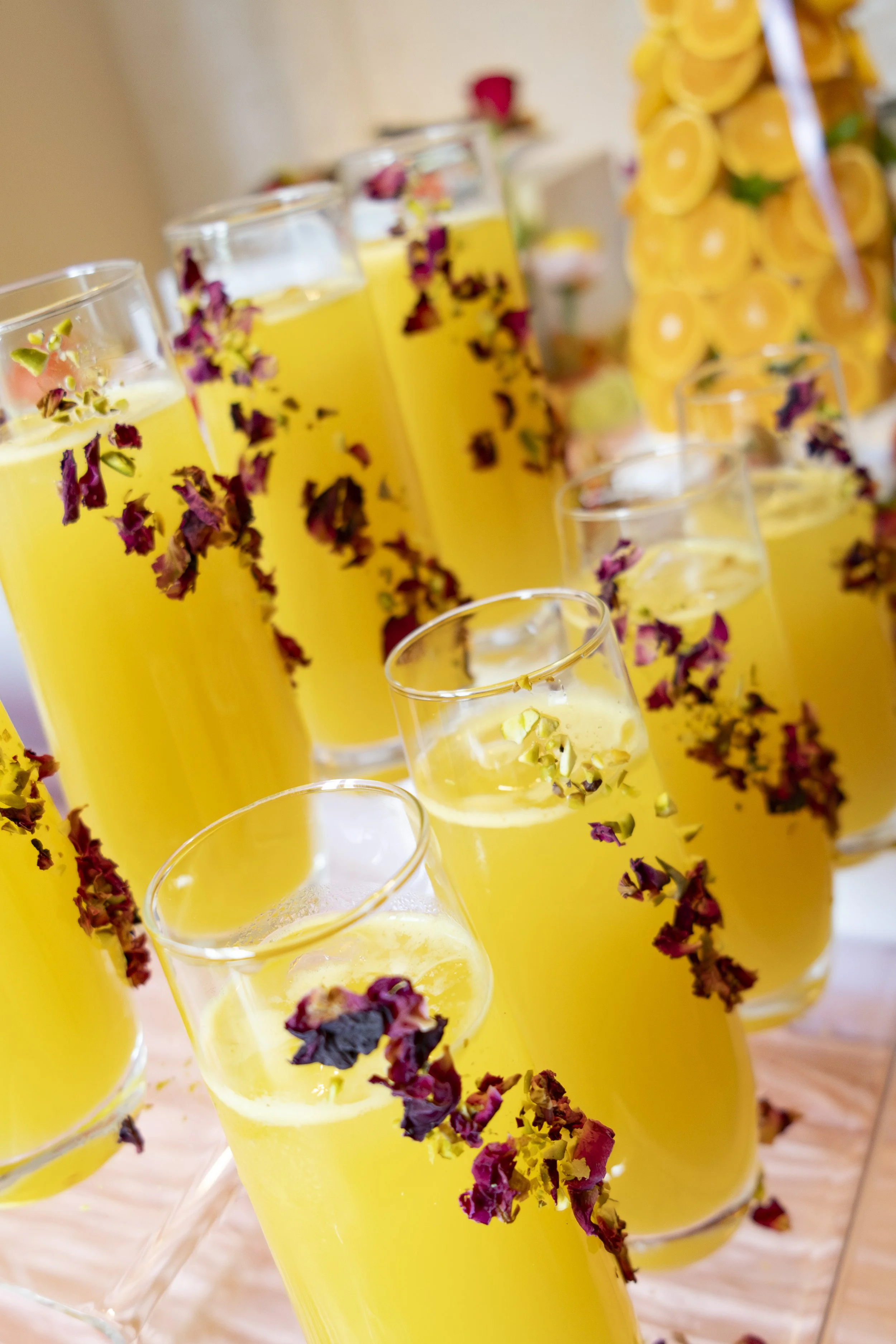 Tall glasses filled with yellow drink, garnished with dried rose petals, on a table with a blurred background of stacked yellow lemonades.
