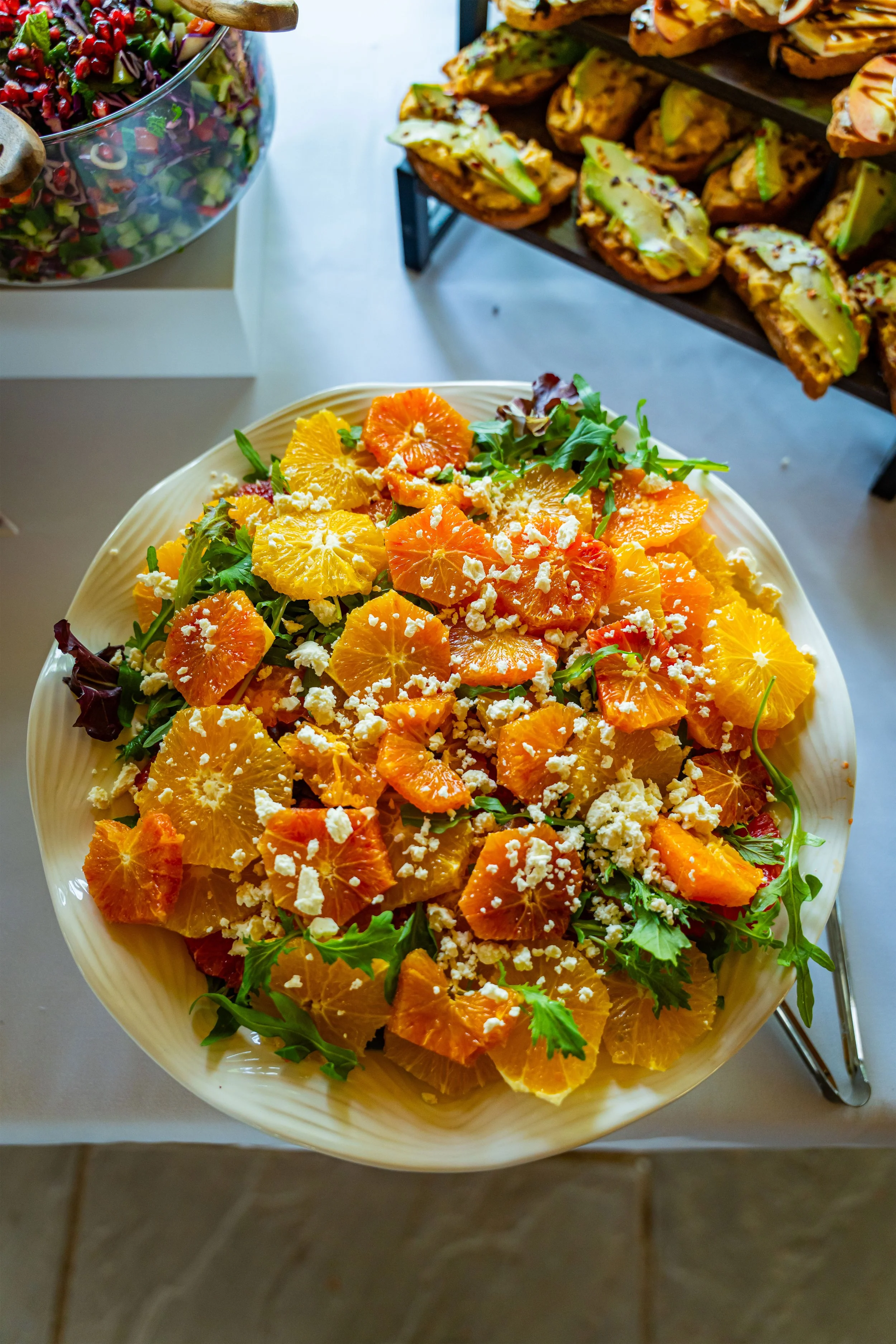 A bowl of mixed salad with orange slices, crumbled cheese, and greens. In the background, there are sandwiches with avocado and other toppings on a wooden tray.