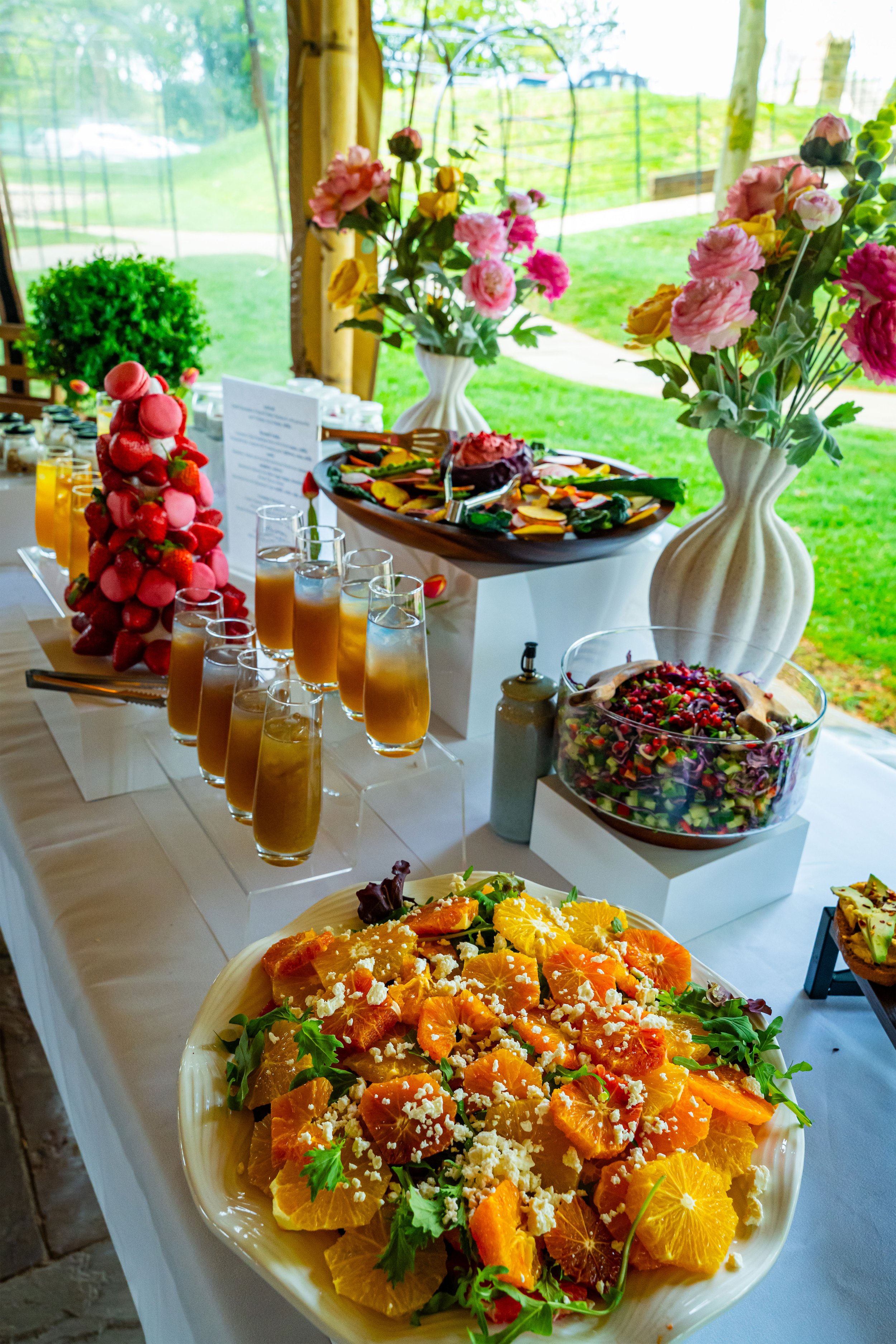 A buffet table with colorful fruit salads, drinks, and floral arrangements in vases, set outdoors under a tent.
