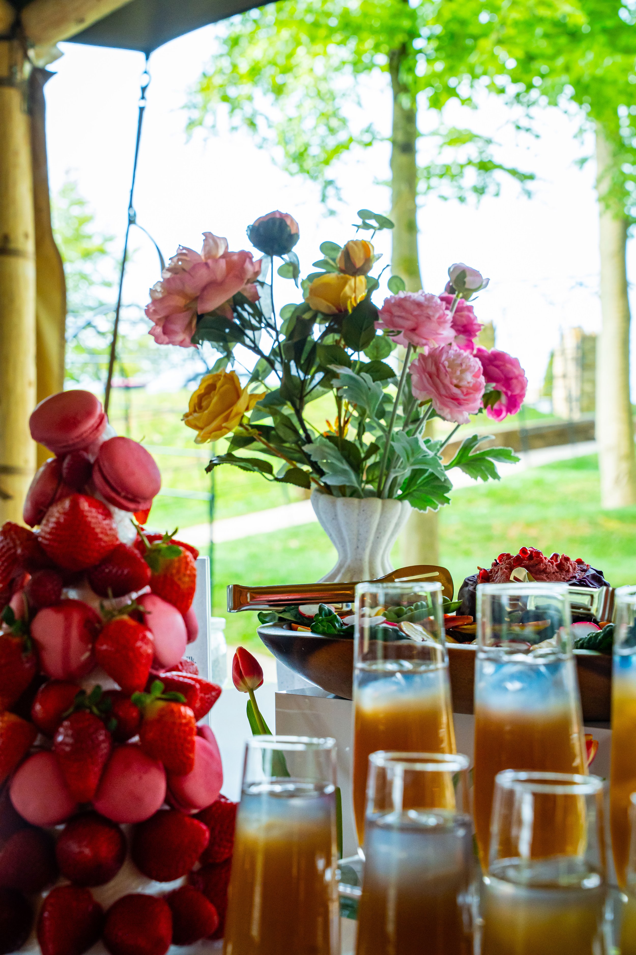 A table with a bouquet of pink and yellow flowers in a white vase, glasses of orange and yellow drinks, a pink macaron tower, strawberries, and other desserts, with green trees and a bright sky visible through the window.