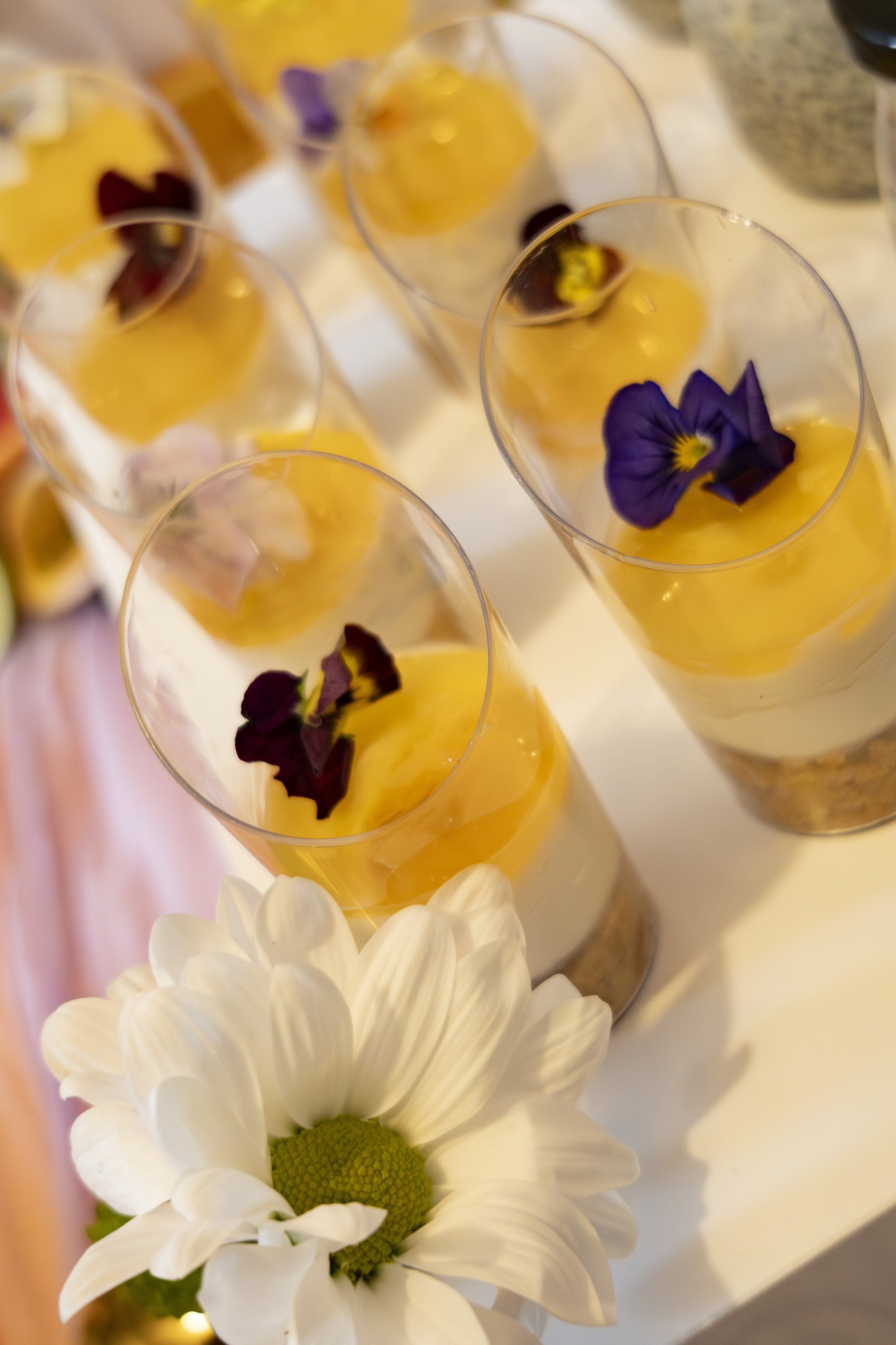 Multiple transparent glasses filled with a yellow-orange beverage, garnished with purple flower petals, set on a white surface. There is a white daisy flower in the foreground.