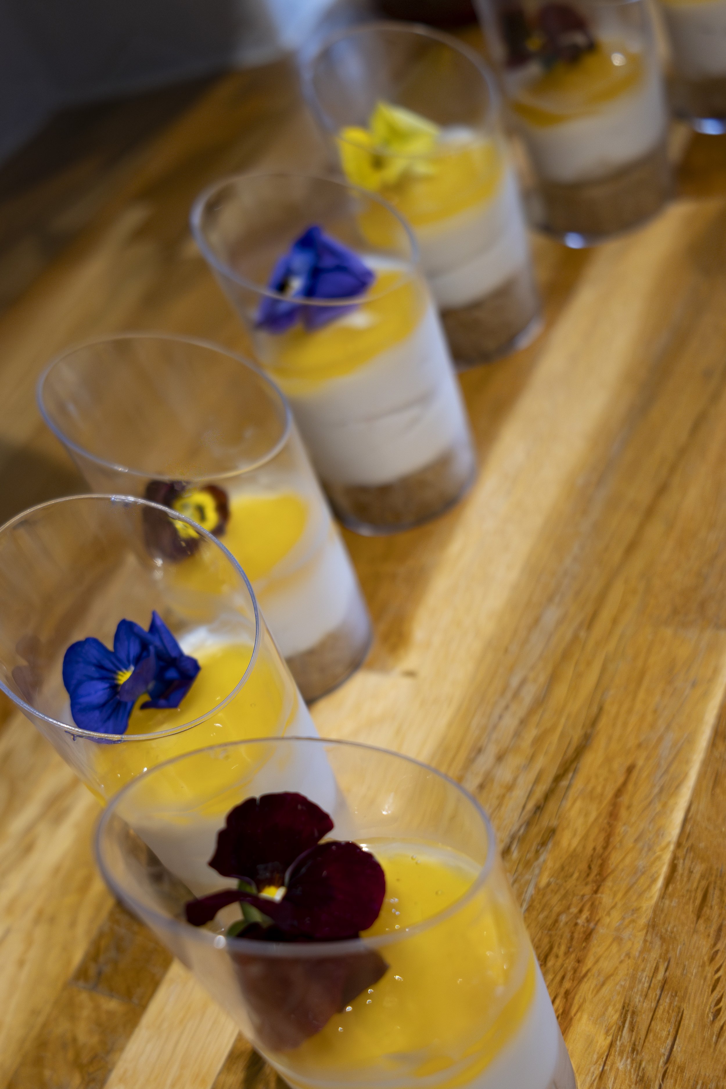 A row of clear glasses filled with a dessert topped with edible flowers and berries, sitting on a wooden surface.