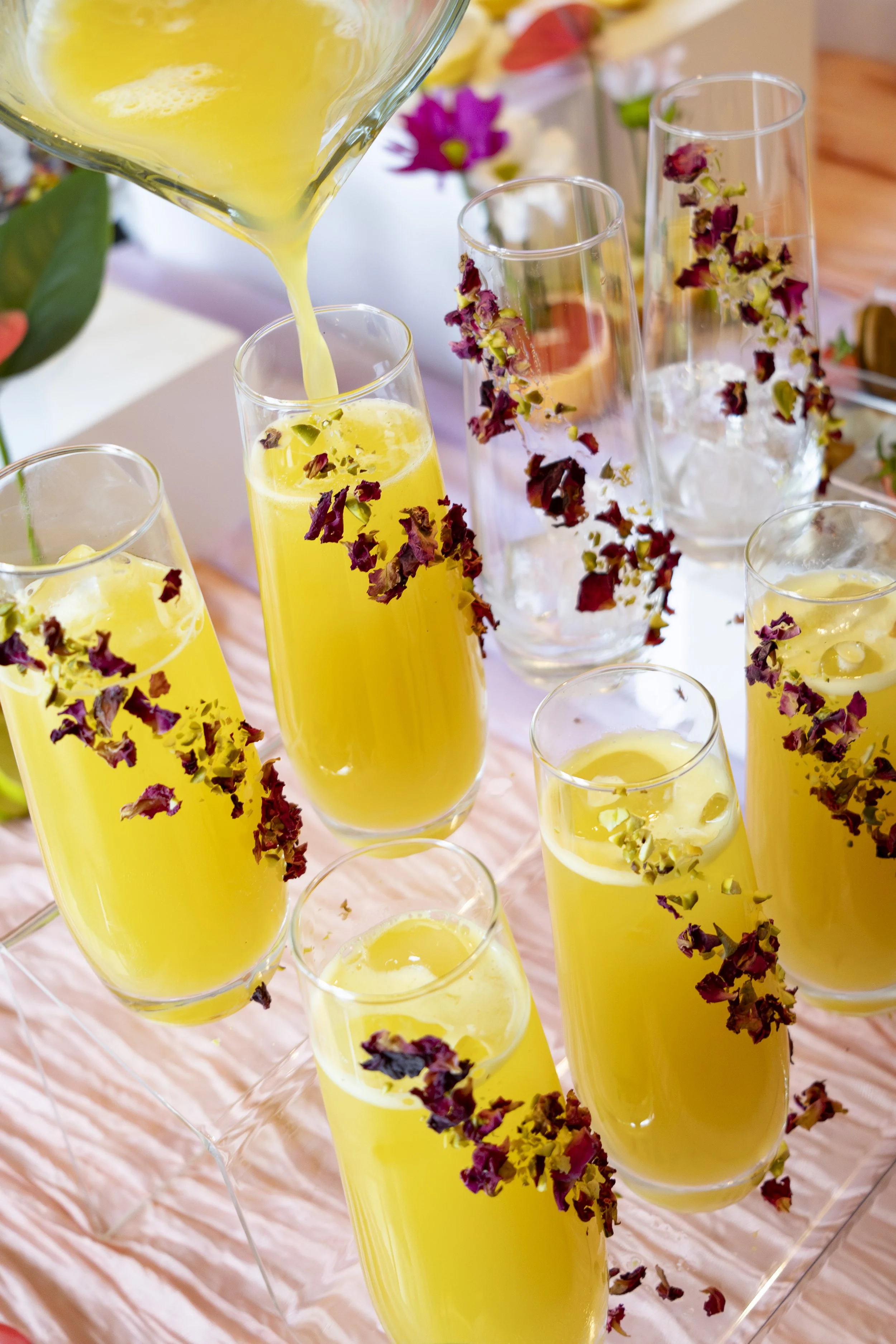 Tall champagne flutes filled with a yellow beverage, decorated with dried flower petals, and ice cubes. A pitcher of yellow liquid is being poured into one of the glasses. In the background, there are pink and purple flowers, and a soft, pink tablecl