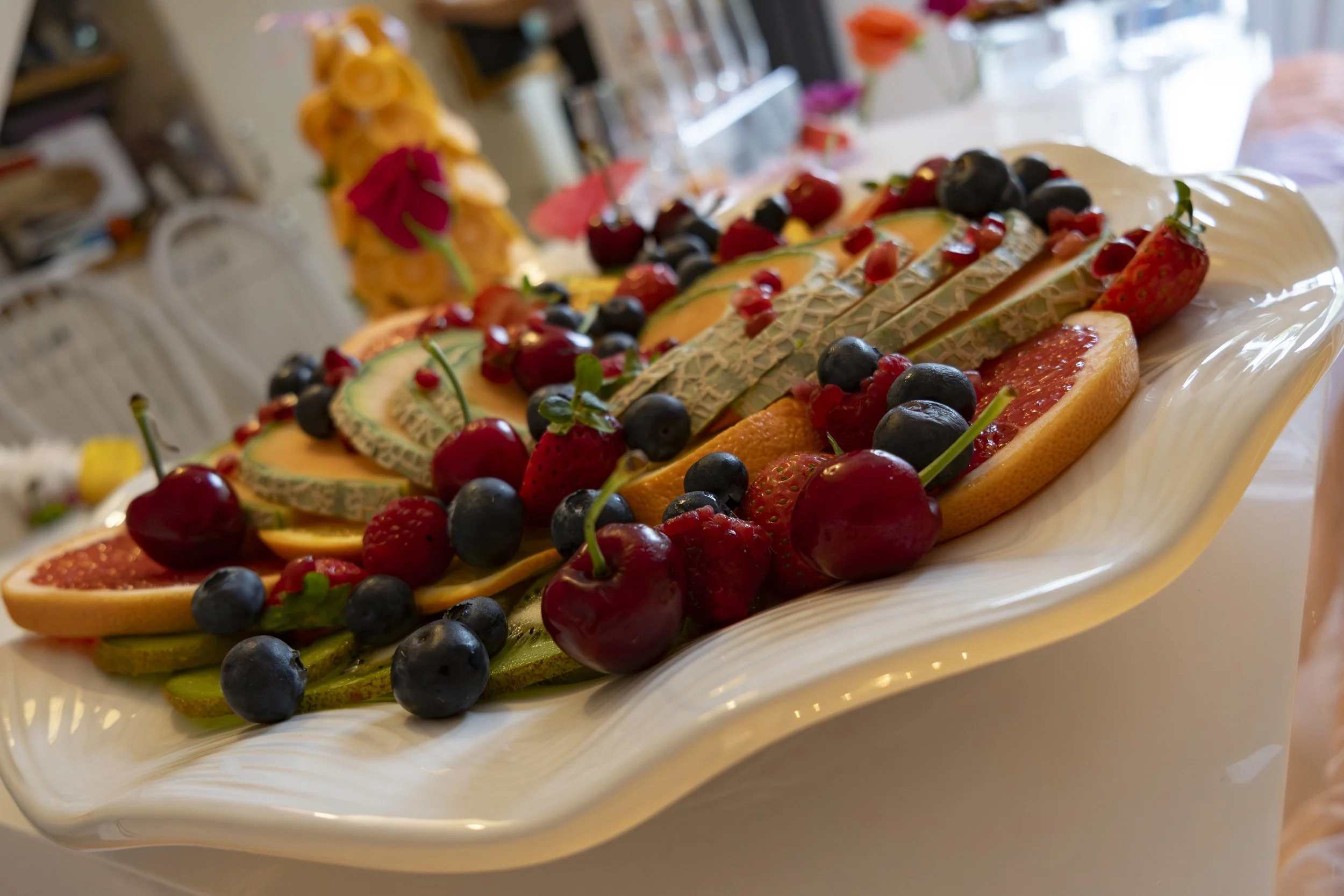 A decorative serving platter filled with an assortment of fresh fruits, including strawberries, blueberries, cherries, and grapefruit slices, with some garnished with small green leaves.