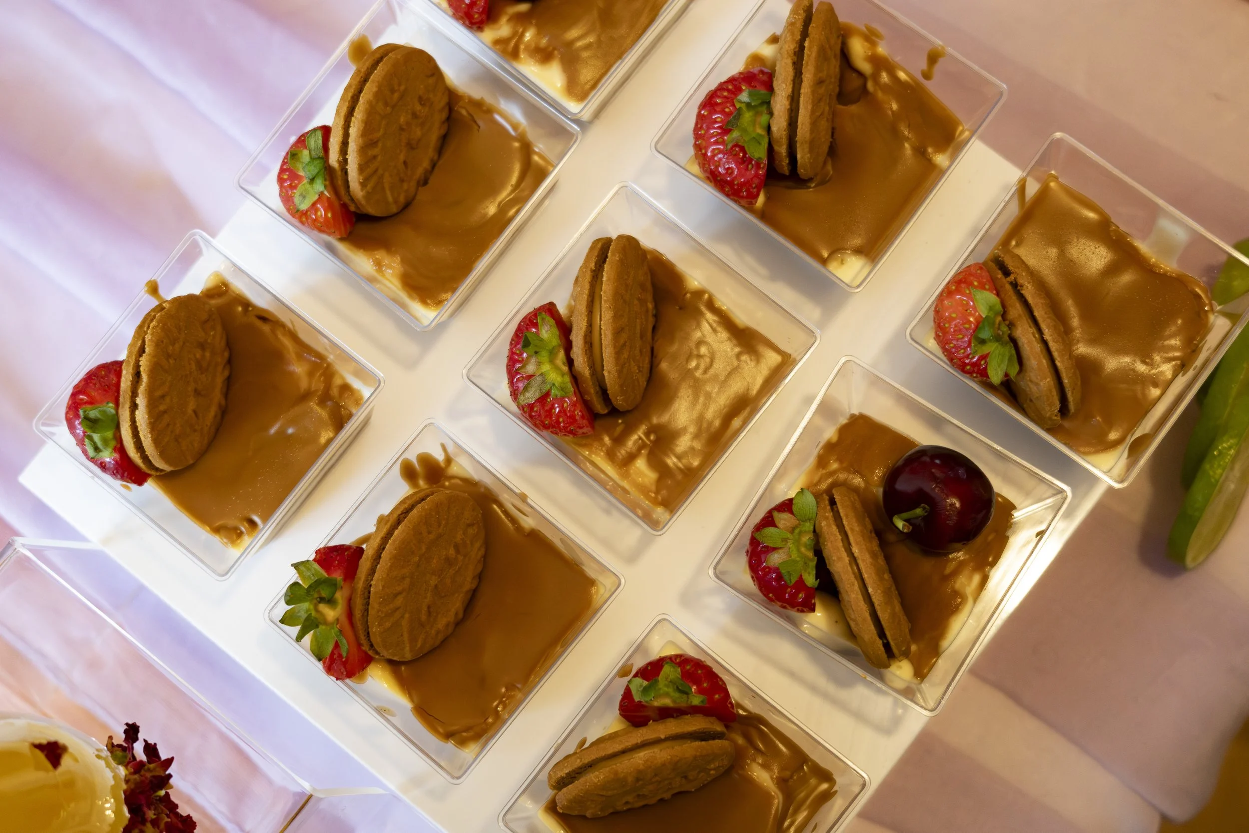 Mini dessert cups filled with caramel sauce, topped with a cookie and a strawberry, arranged in a grid on a white tray.