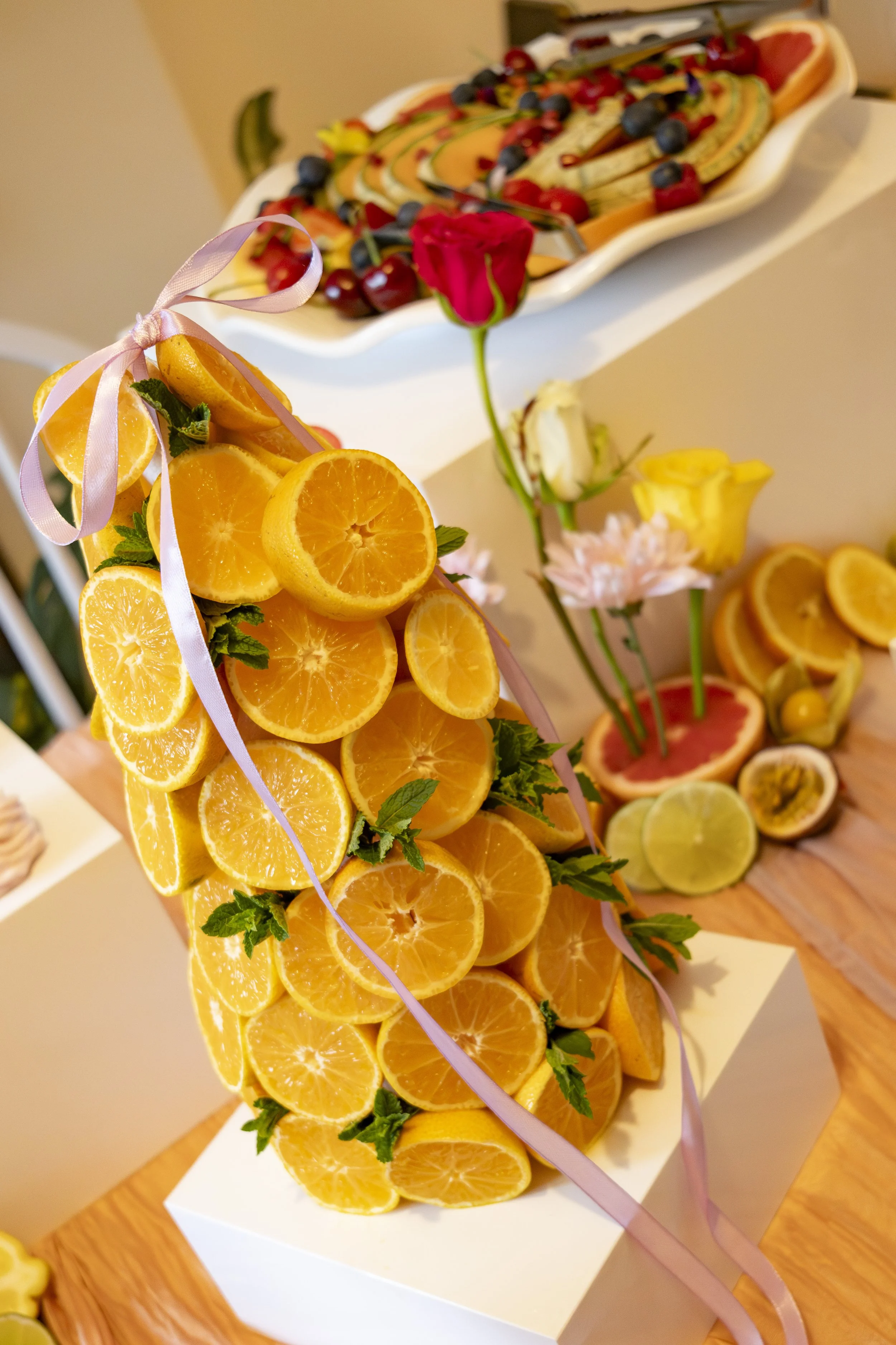 A floral arrangement made of orange slices, decorated with a pink ribbon, with a background of fruit and floral displays including sliced lemons, limes, and a pink flower.