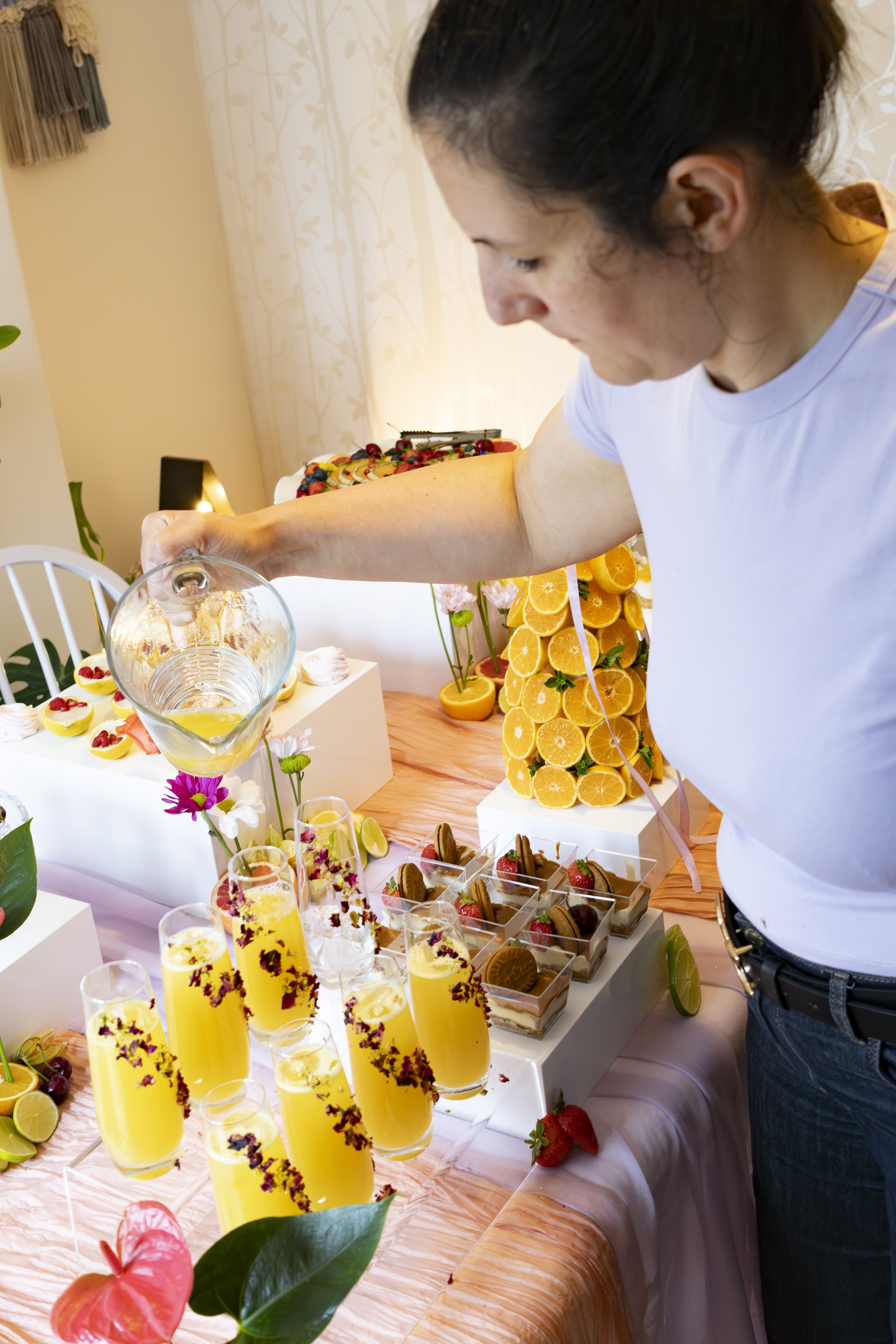 A woman with dark hair tied back is pouring a yellow drink into a glass at a table with an assortment of desserts, fruits, and flowers, including orange slices, strawberries, and macarons, in a brightly decorated room.