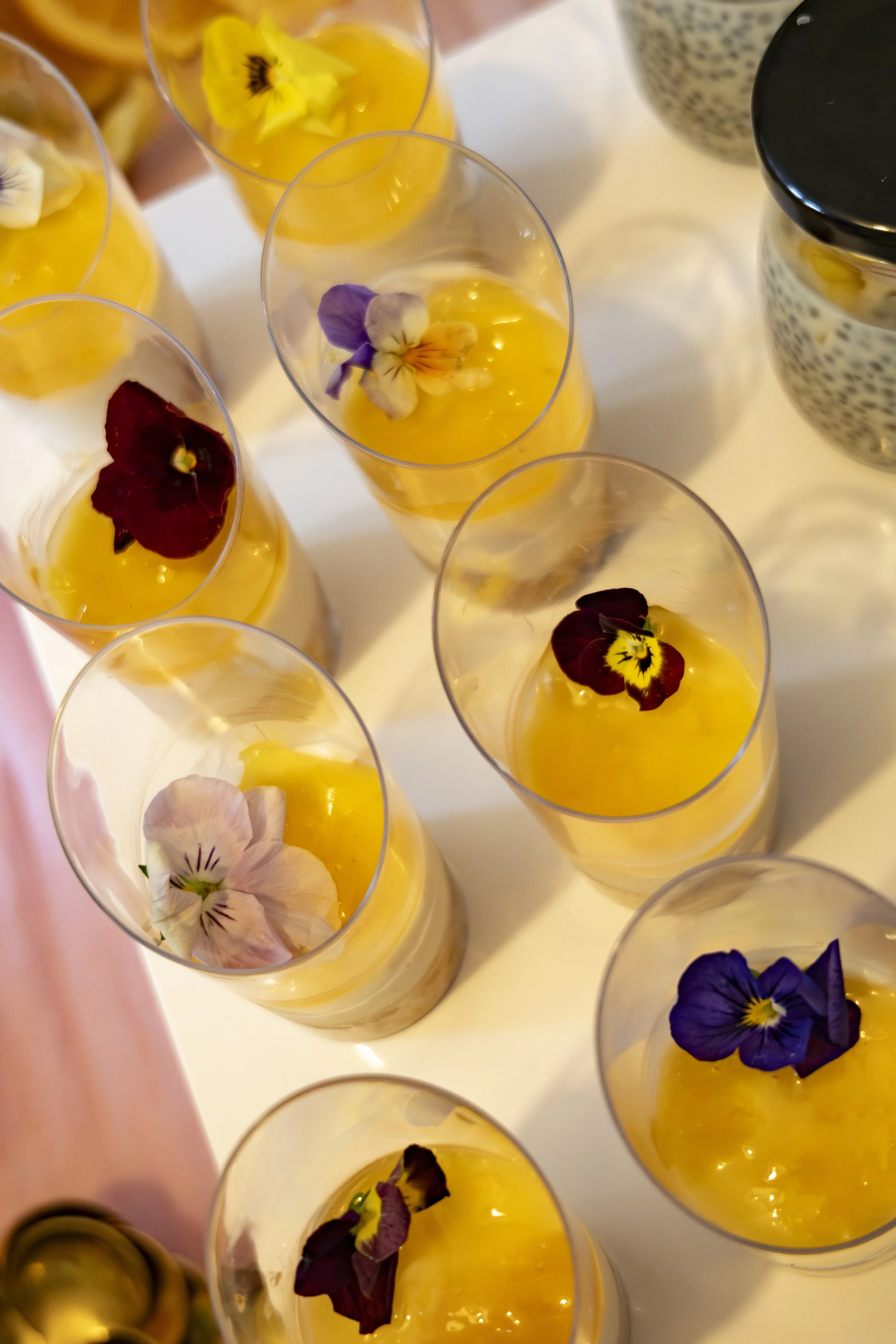 Multiple small glasses filled with a yellow dessert topped with edible flower petals, arranged on a white surface.